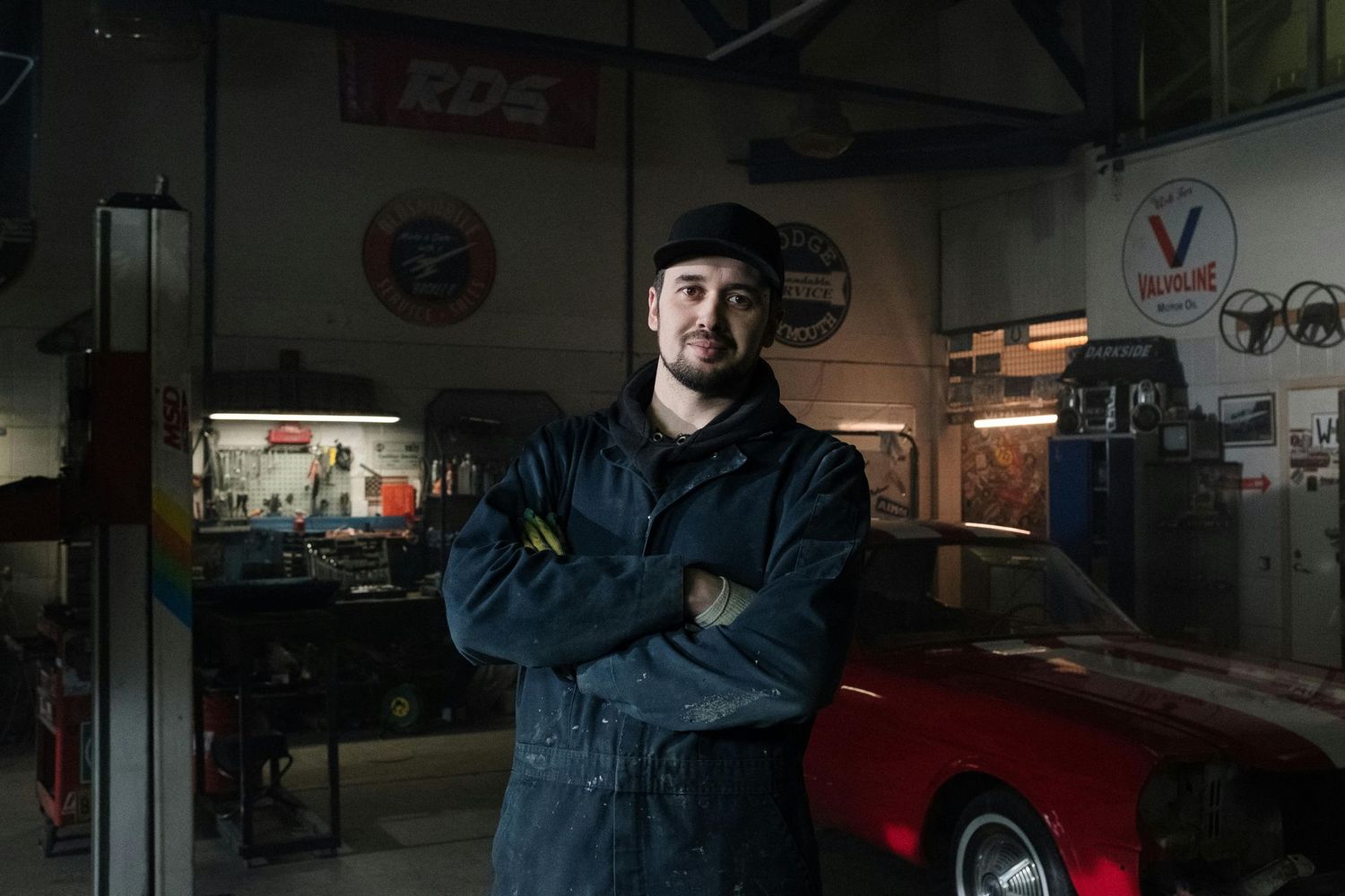Mechanic in blue overalls smiles, arms crossed, in a garage with a red car.