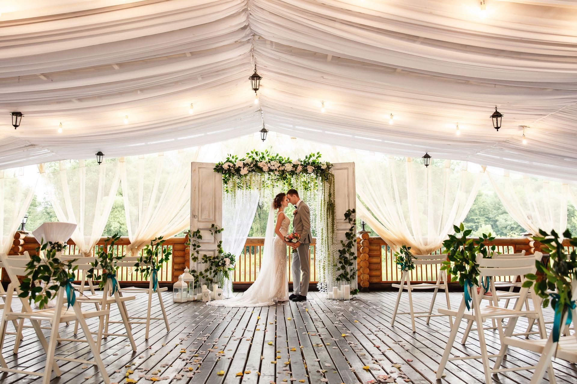Wedding ceremony: Couple kissing under floral arch, wooden deck, draped white fabric ceiling.