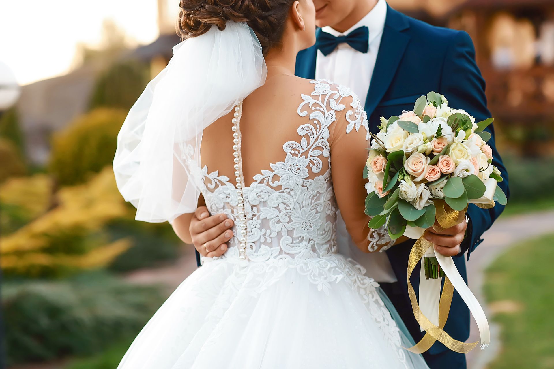 Bride in white lace dress and veil embraces groom in blue suit, holding bouquet, outdoor setting.