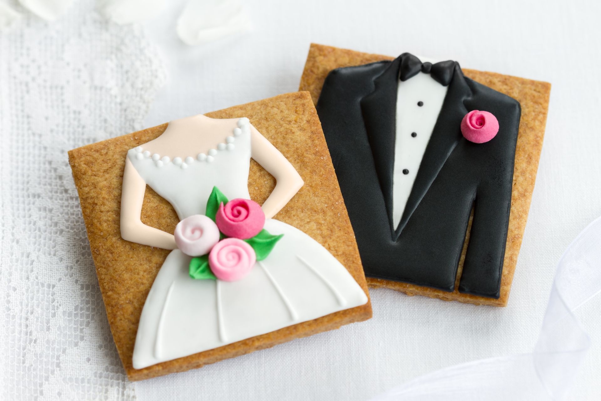 Wedding cookies shaped like a bride's dress with flowers and a groom's tuxedo, on a white lace surface.