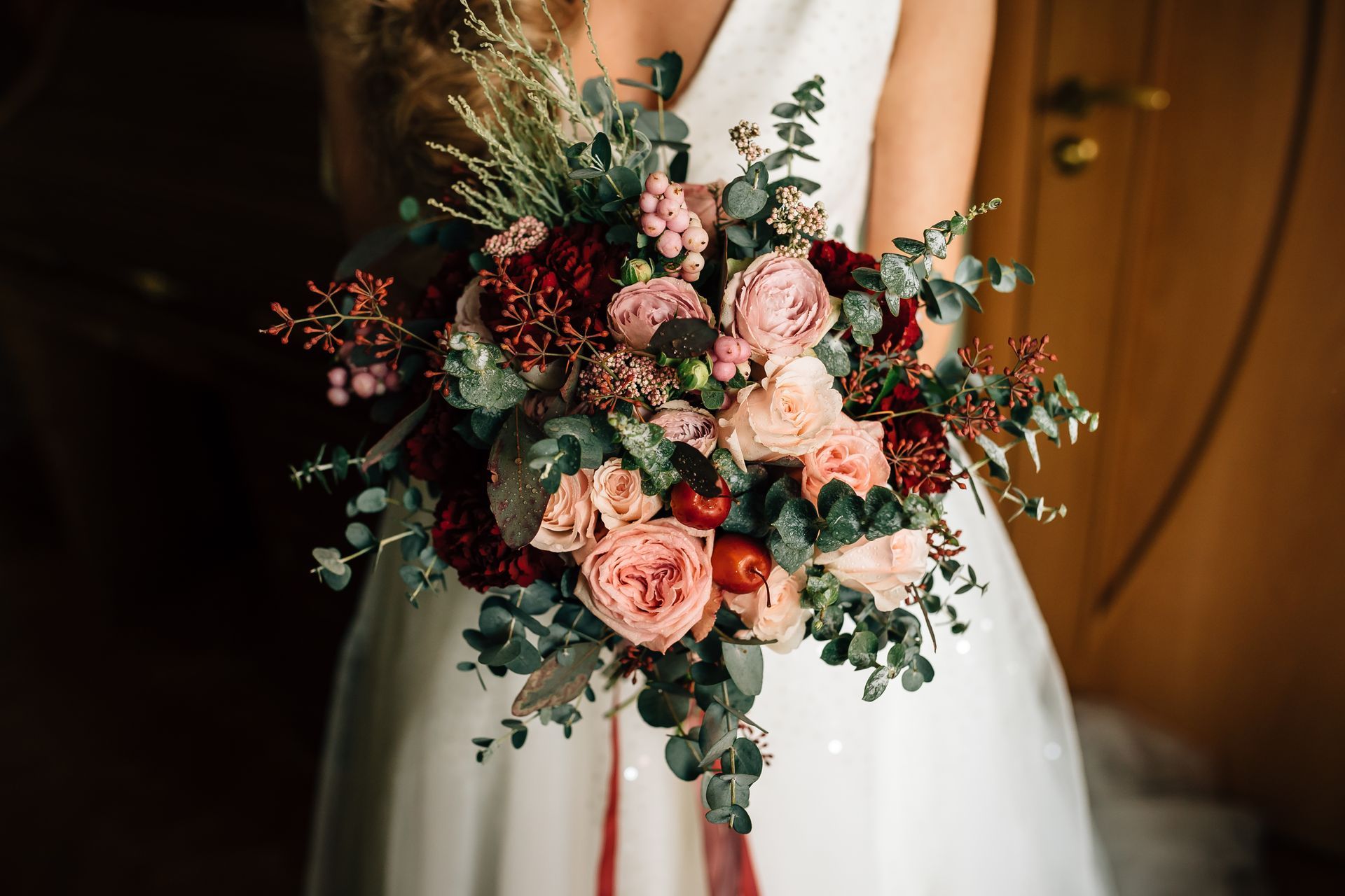 Bride in a white dress holding a bouquet of red and pink flowers, with green foliage, near a wooden door.