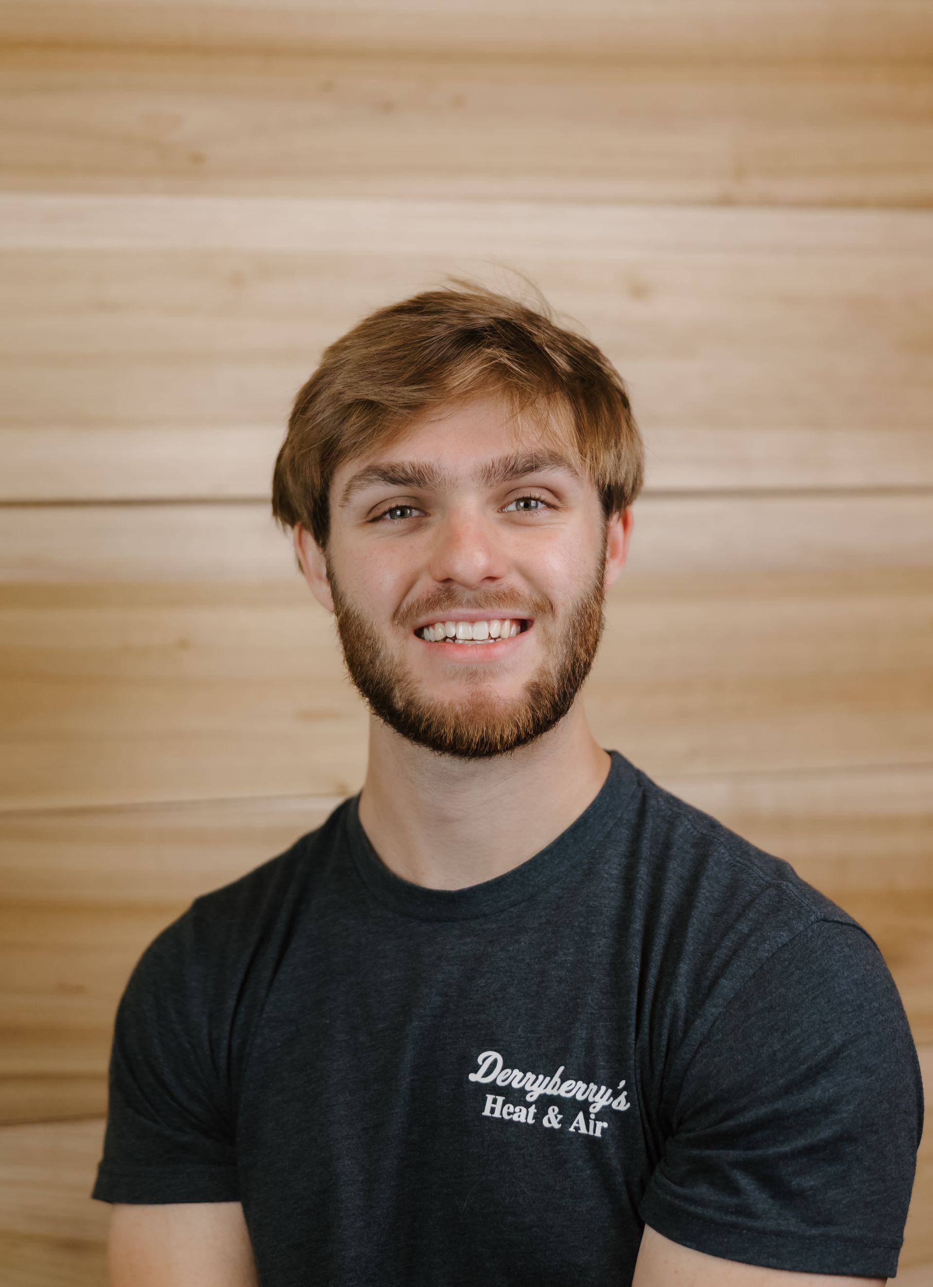 Man smiles at the camera, with light brown hair and a beard, wearing a gray t-shirt with a logo. Wooden background.