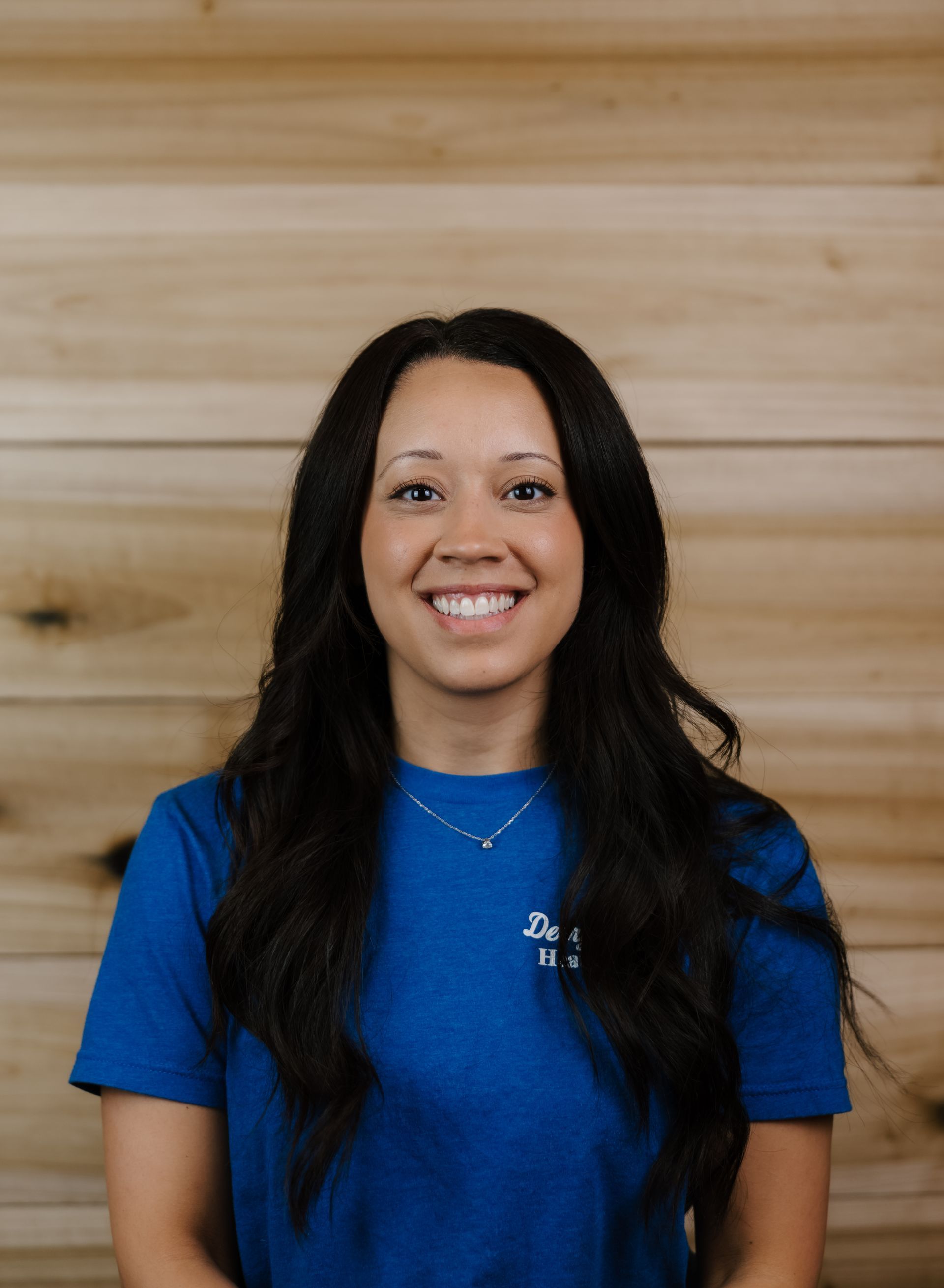 Woman with long dark hair, smiling, wearing a blue shirt, standing in front of wood paneling.