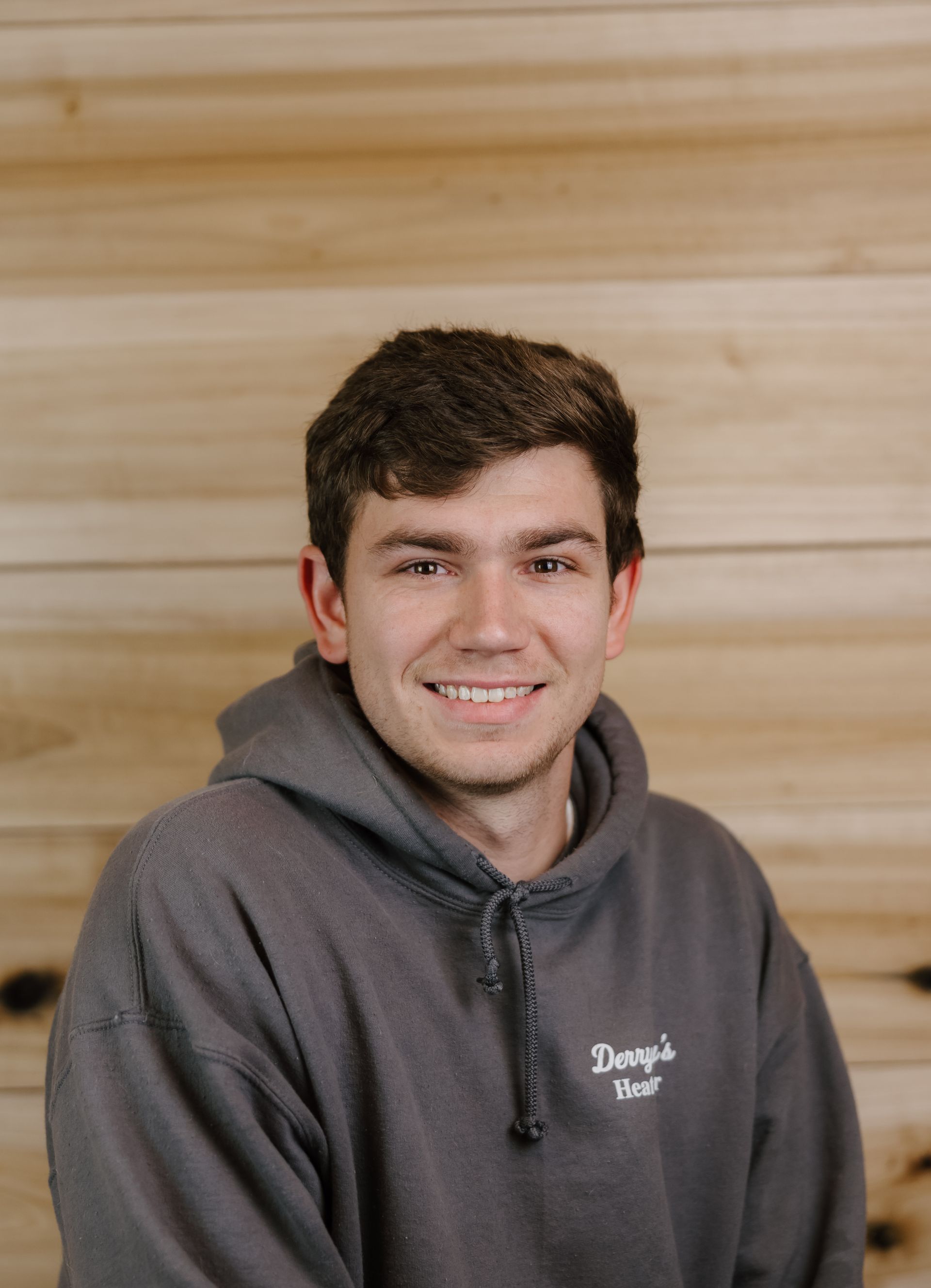 Man smiling, wearing a gray hooded sweatshirt, standing in front of a wood panel background.