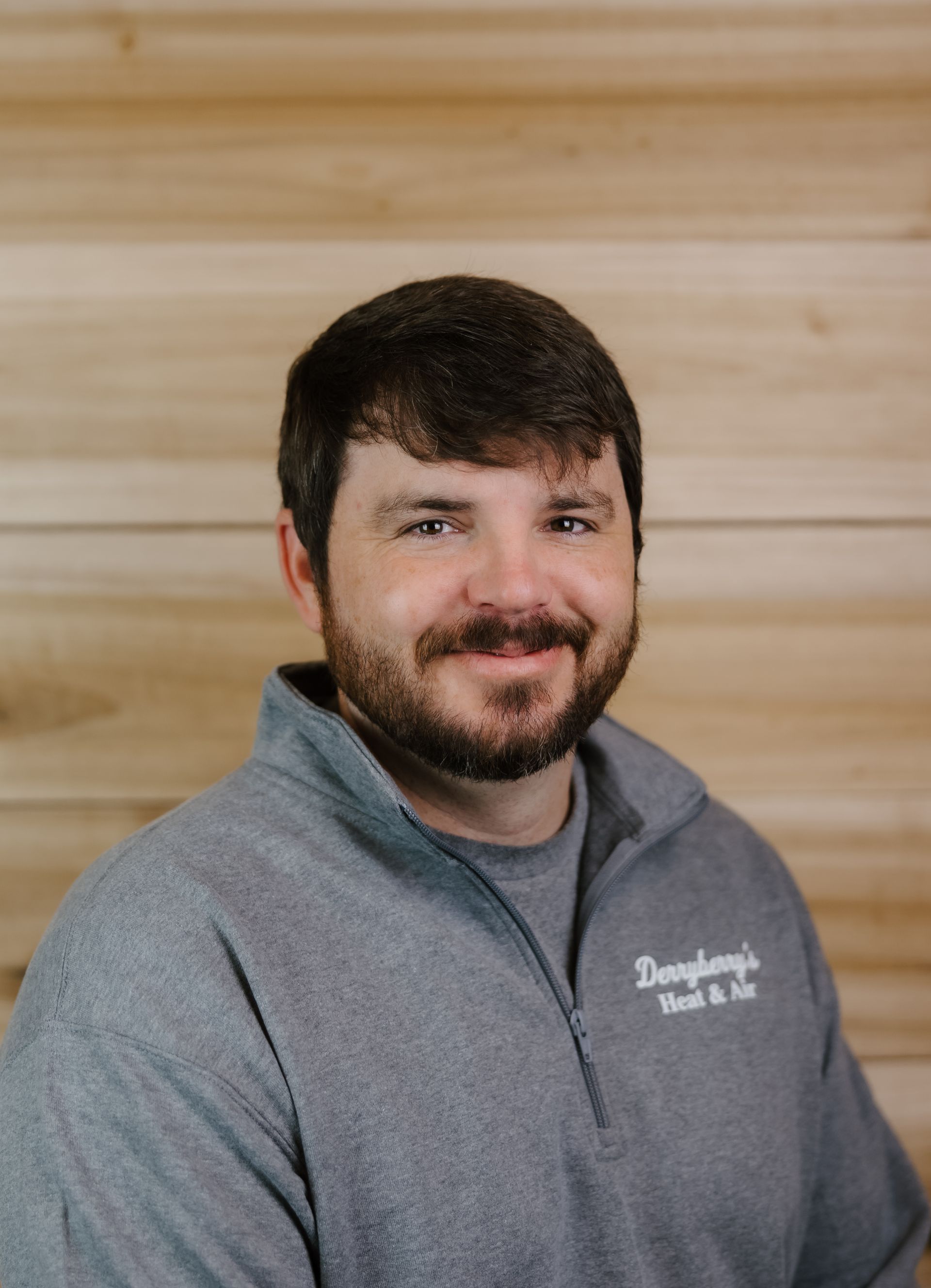 Man wearing a gray sweater with logo, smiling, standing in front of wood paneling.