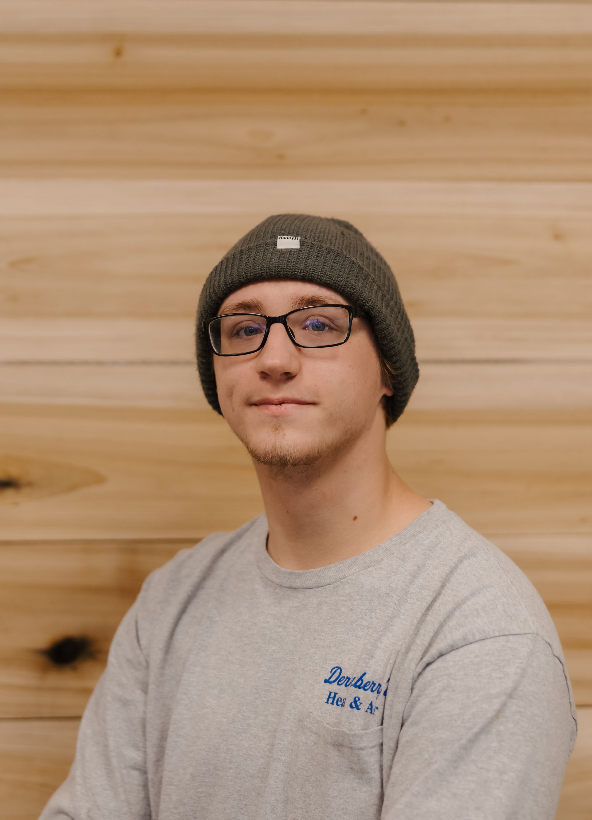 Man with glasses and a gray beanie smiles, in front of a wooden wall.