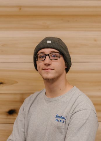 Man with glasses and a gray beanie smiles, in front of a wooden wall.