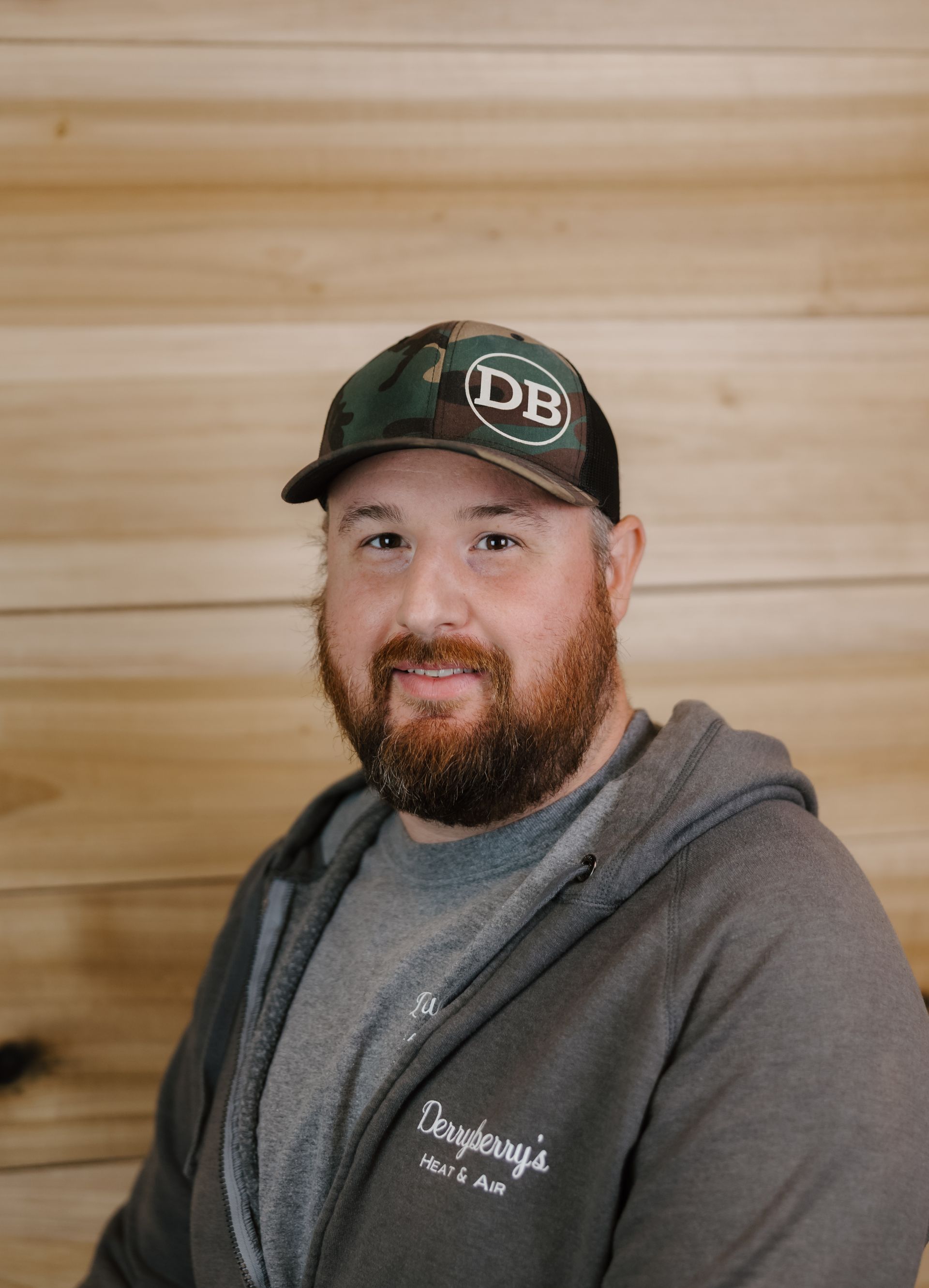 Man wearing camouflage cap and gray hoodie, smiling, in front of a wood background.