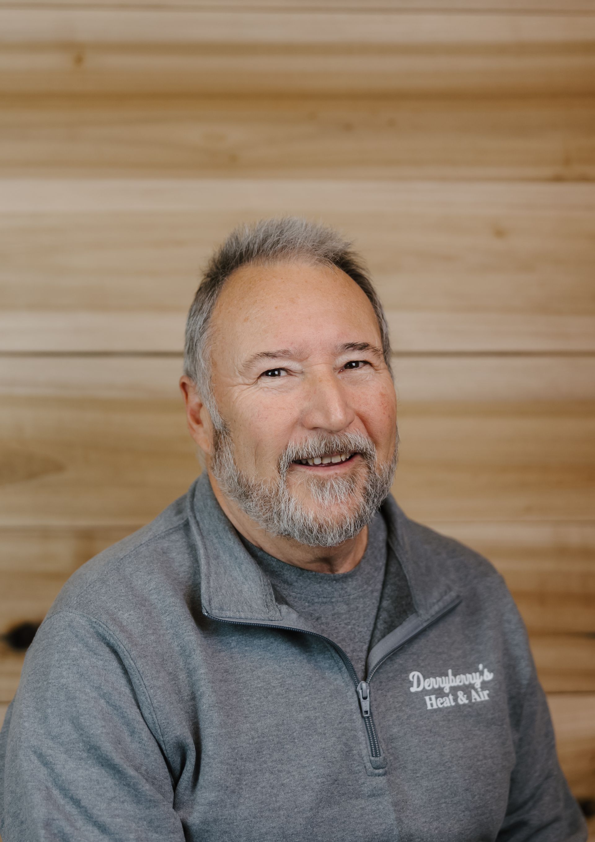 Man with a gray beard and gray sweater in front of a wooden wall. Sweater says 