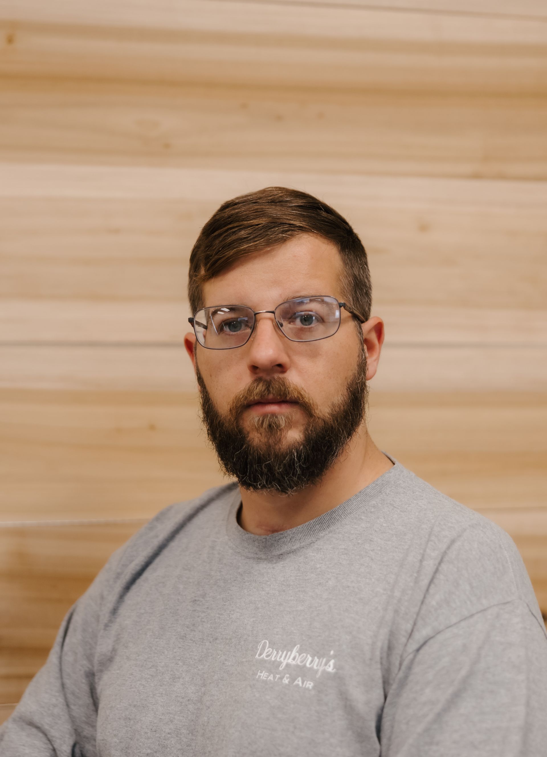 Man with glasses and beard, wearing a gray sweatshirt, in front of a wood-paneled wall.