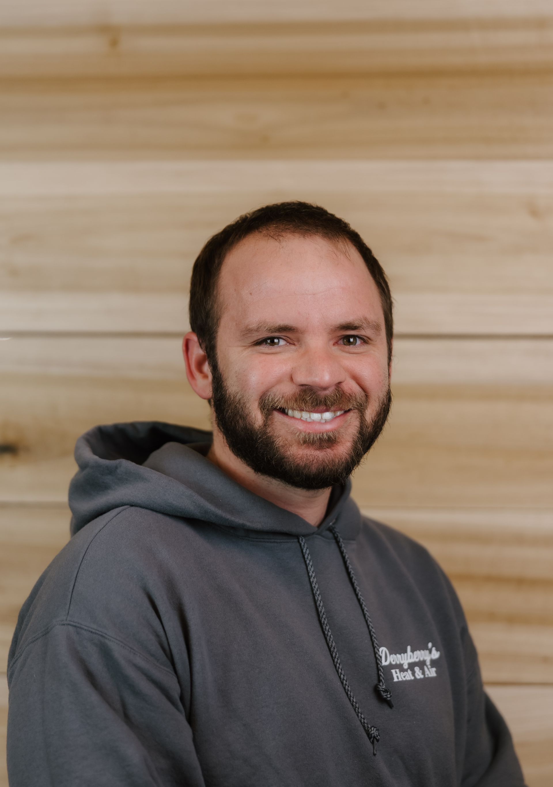 Man in gray hoodie smiles, standing against wooden wall.