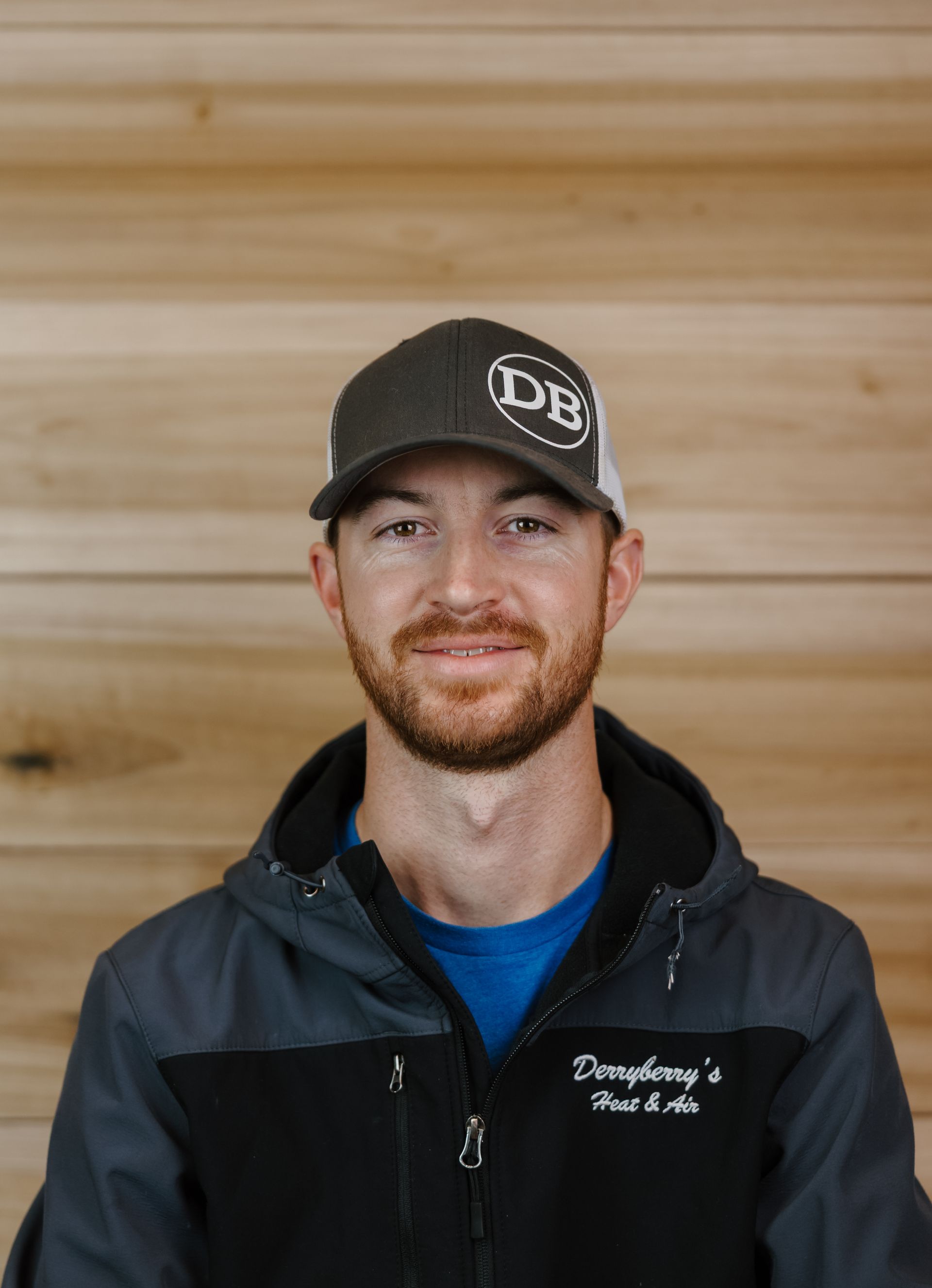 Man wearing a cap and jacket in front of a wood background. Smiling.