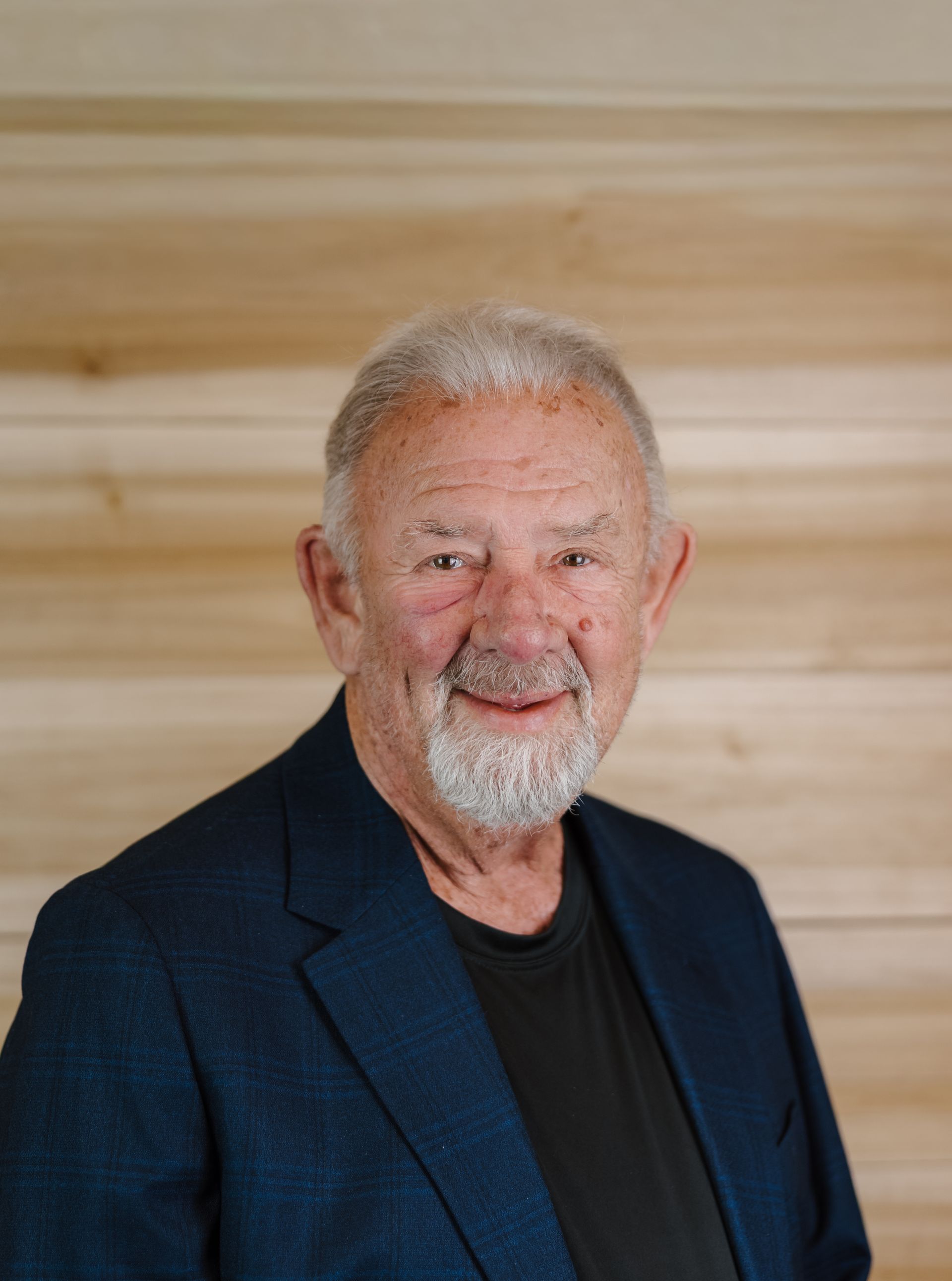 Older man with a gray beard and hair, wearing a navy blazer and black shirt, smiles.