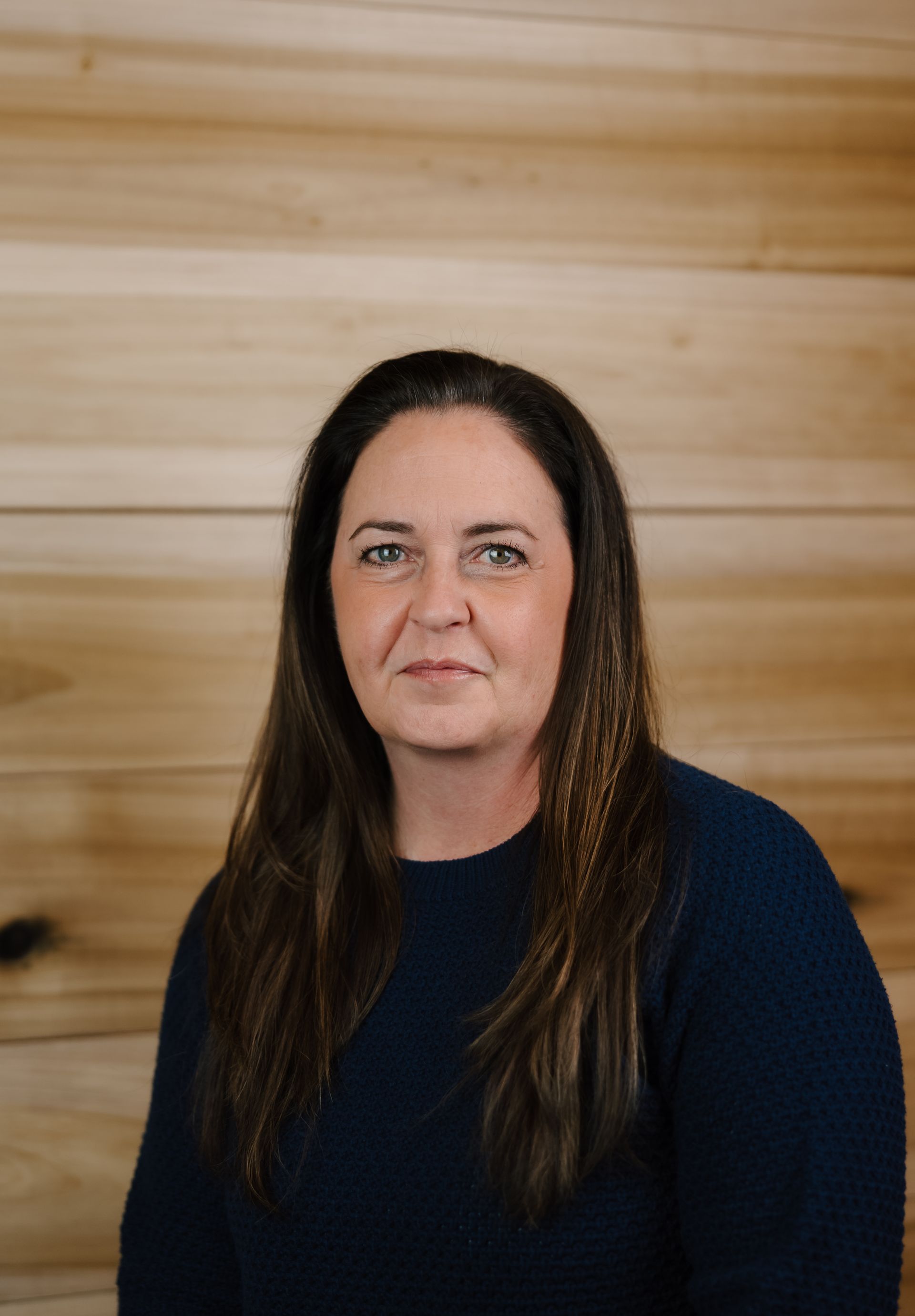 Woman with long brown hair, wearing a dark blue sweater, against a light brown wooden background.