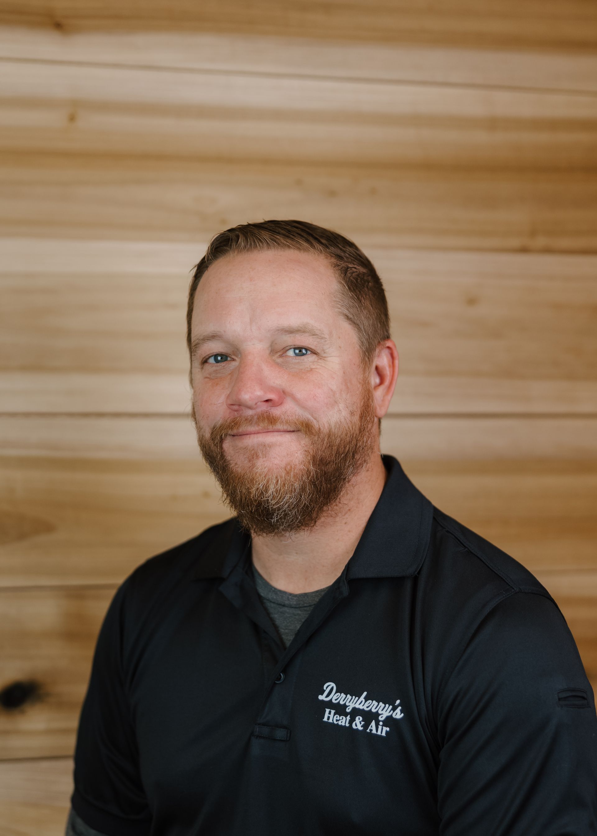 Man with beard wearing black polo shirt smiles in front of a wood background.