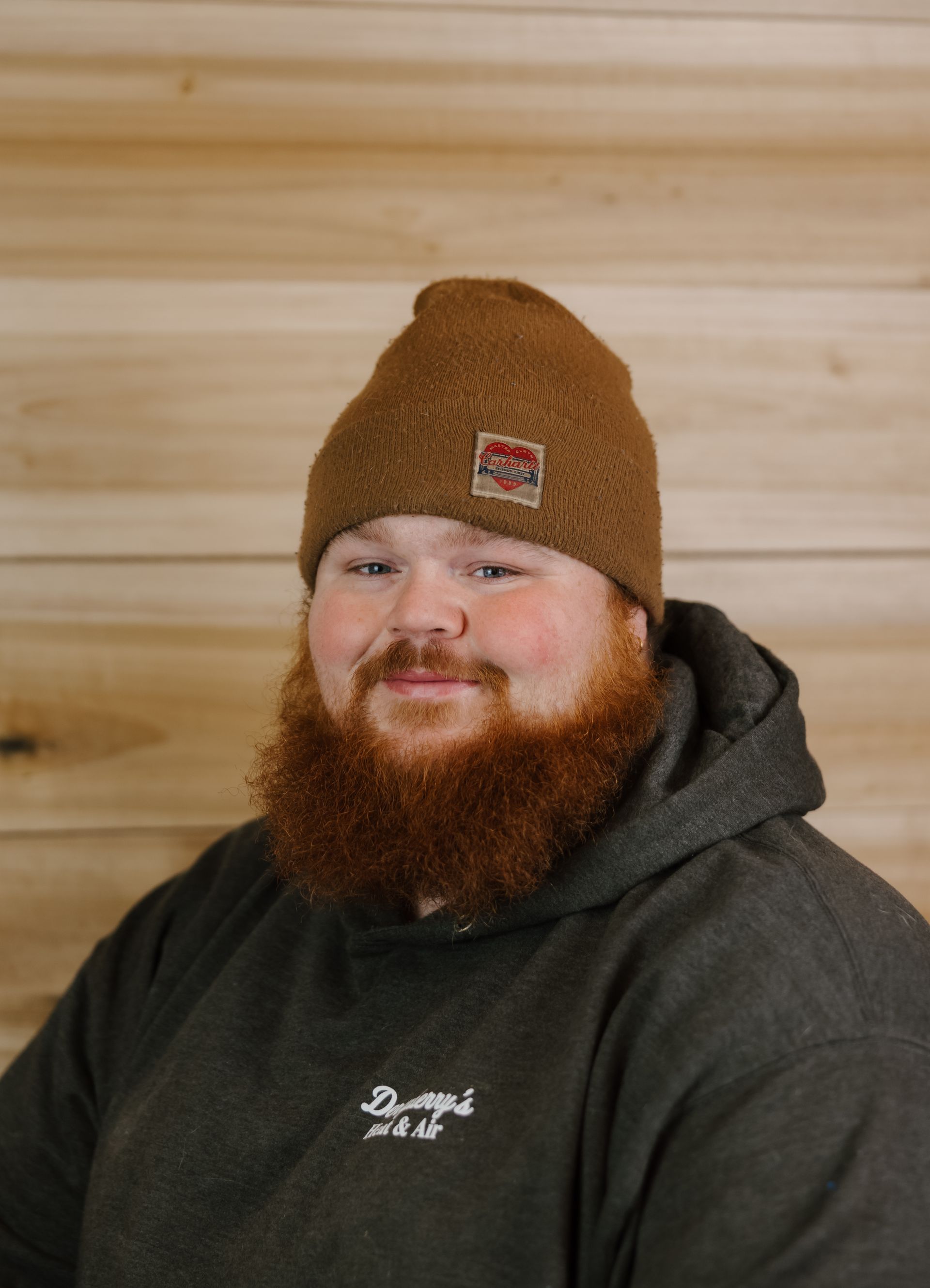 Man with a red beard and brown beanie smiles, wearing a grey hooded sweatshirt in front of wood paneling.
