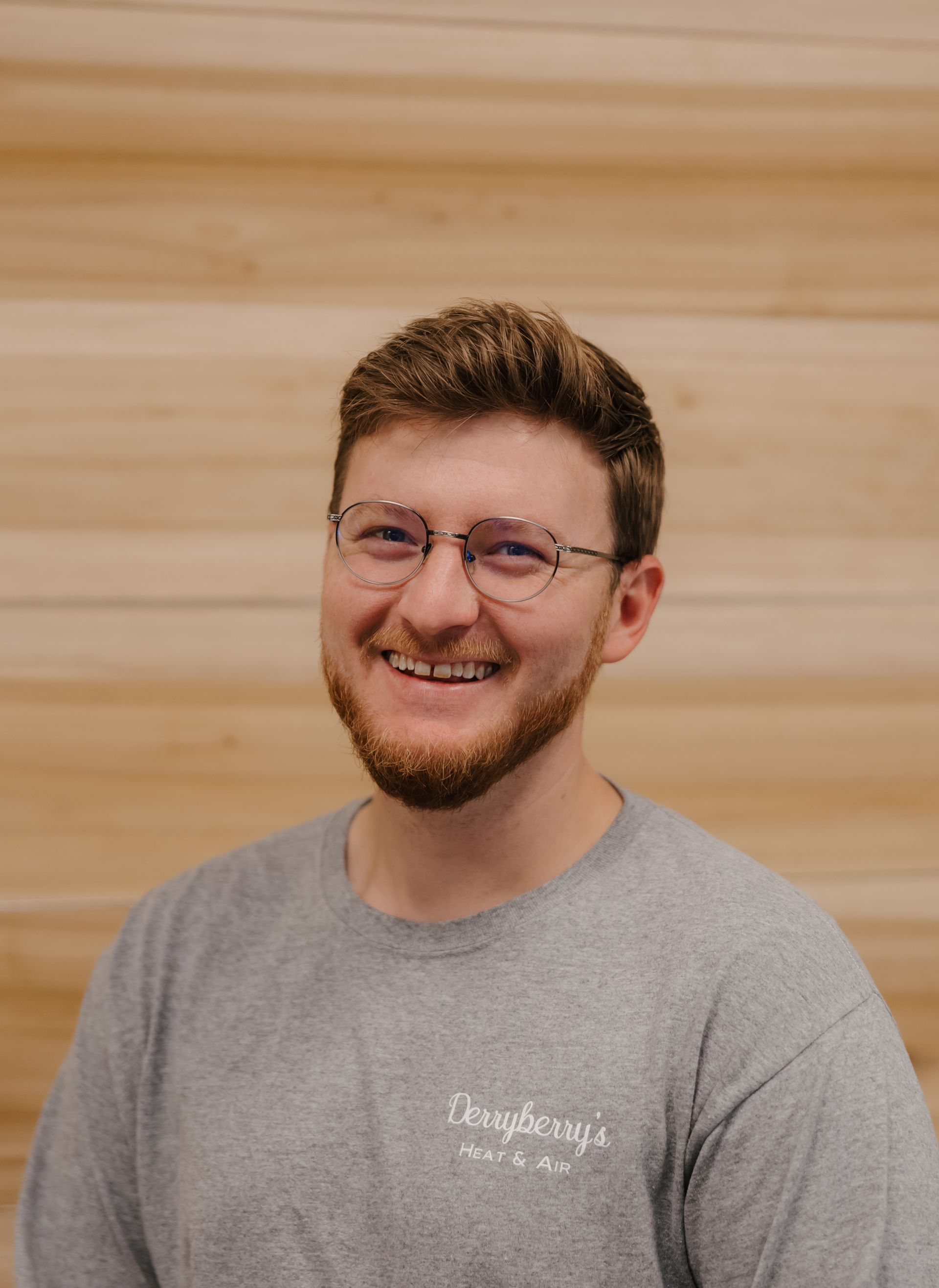 Man with glasses and beard smiles in front of wood-paneled wall, wearing a gray t-shirt.