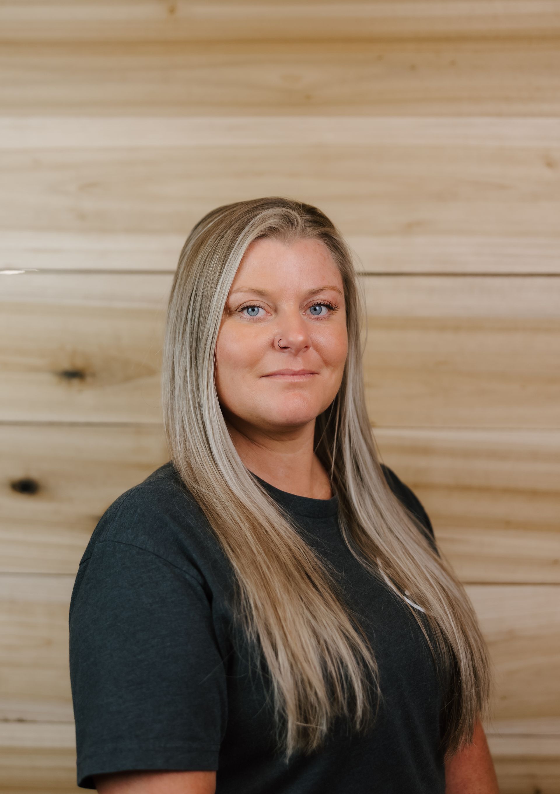 Woman with long blonde hair, wearing a dark green shirt, smiling in front of a wood panel background.