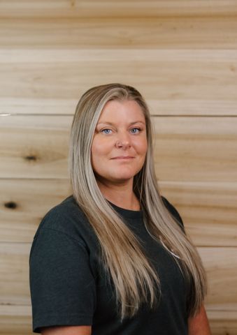 Woman with long blonde hair, wearing a dark green shirt, smiling in front of a wood panel background.
