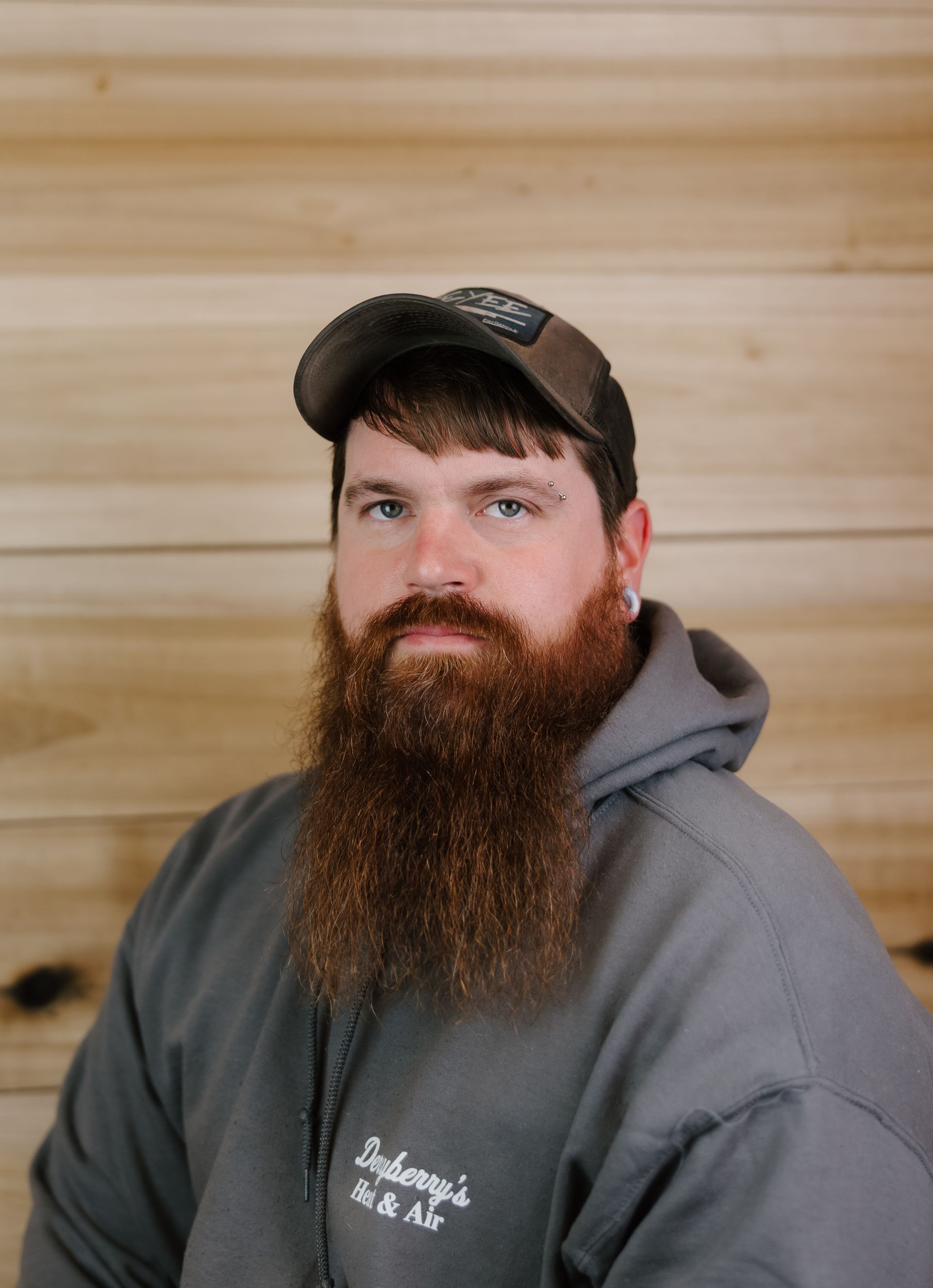 Man with a long beard and a hat wearing a gray hoodie in front of a wood panel background.