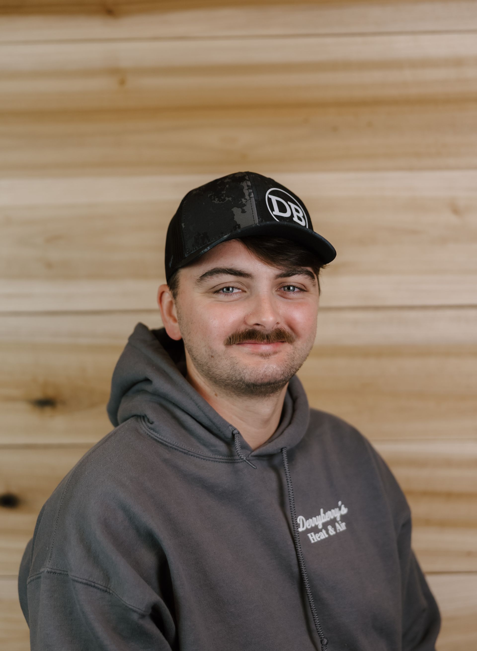 Man with a mustache wearing a black cap and gray hoodie, in front of a wood background.