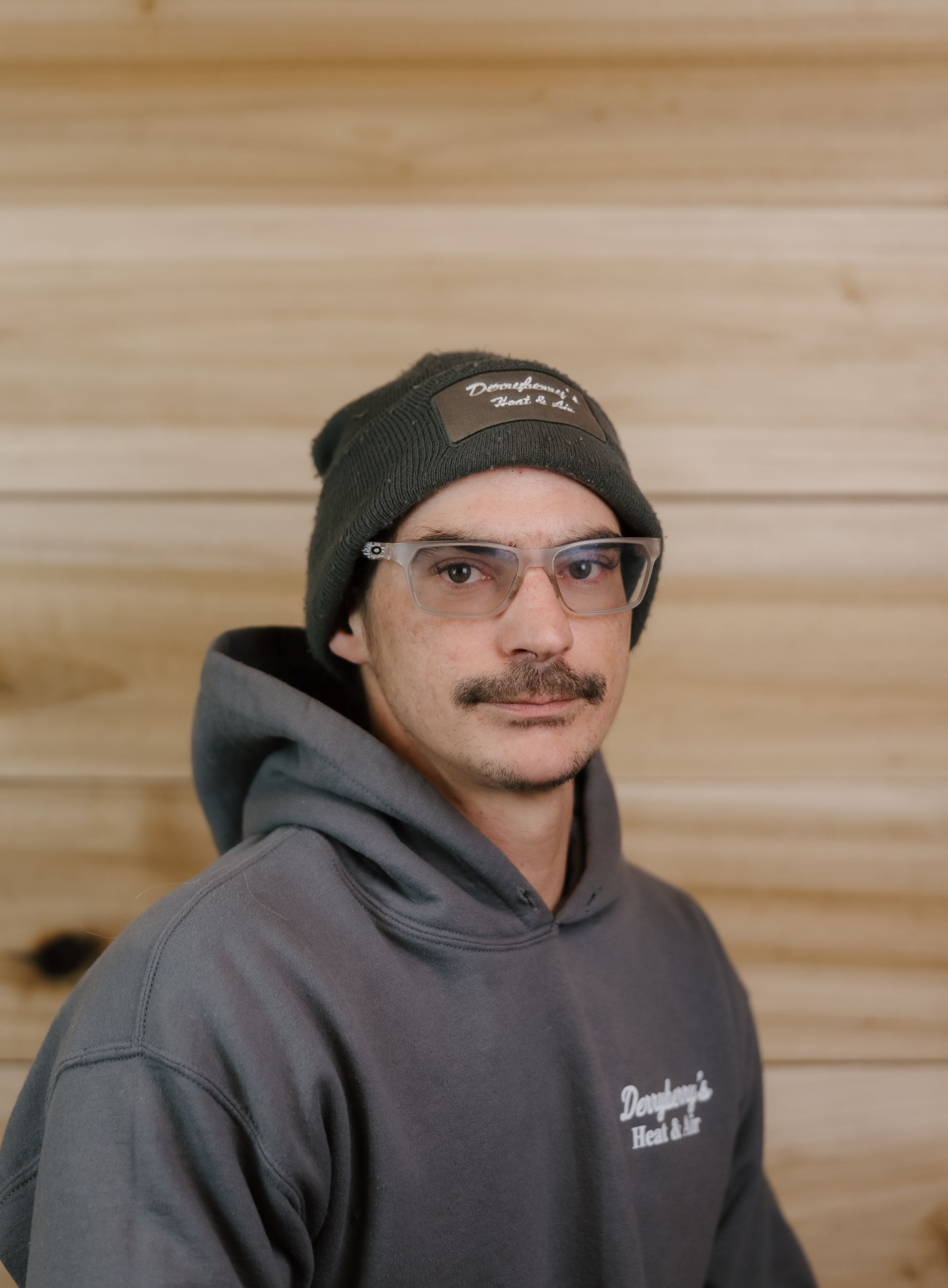 Man with glasses and a beanie, wearing a gray hoodie, in front of a wood background.