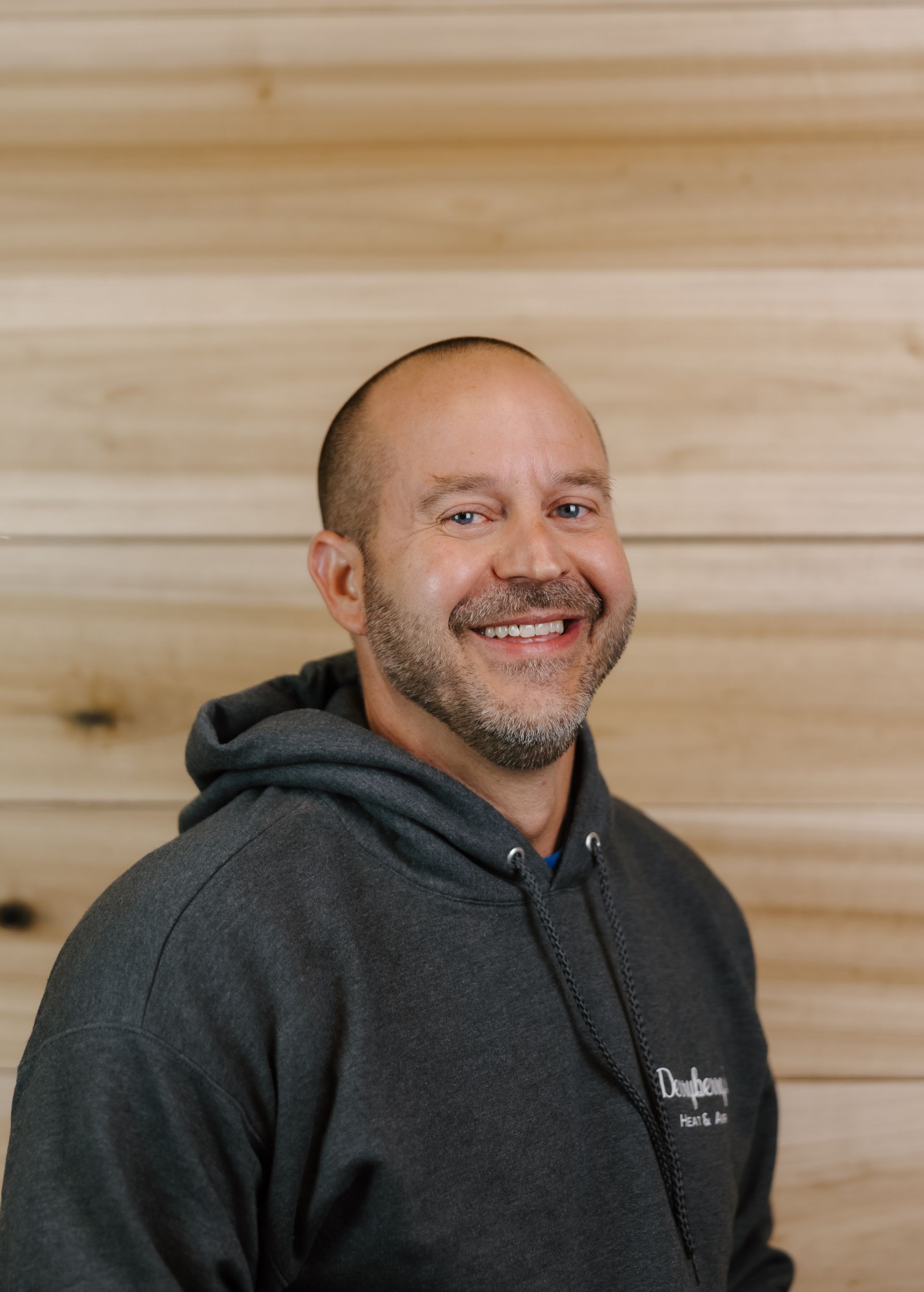 Man in gray hoodie smiles in front of a wood-paneled wall.