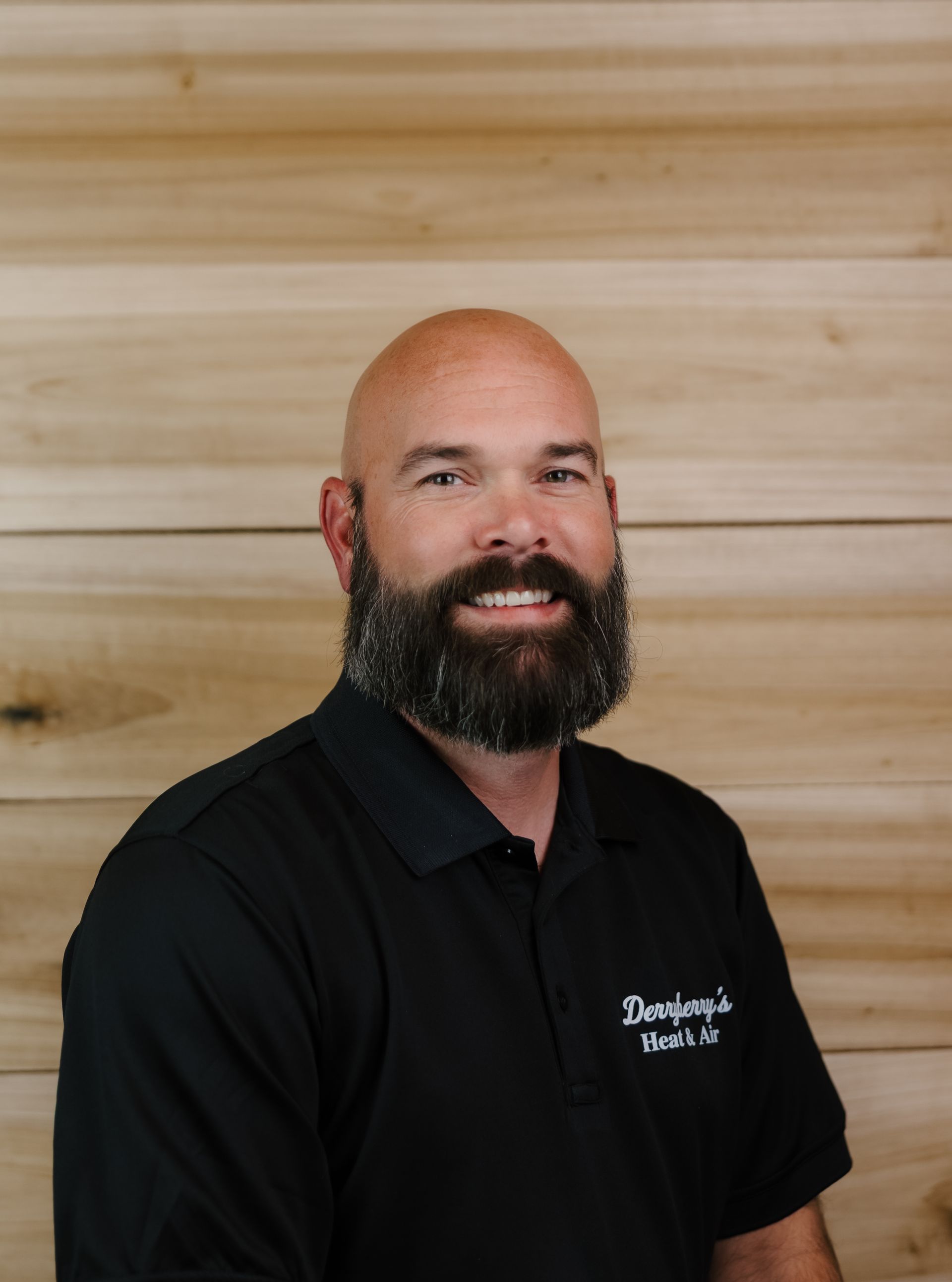 Bald man with a full beard wearing a black polo shirt, smiling in front of a wood-paneled wall.
