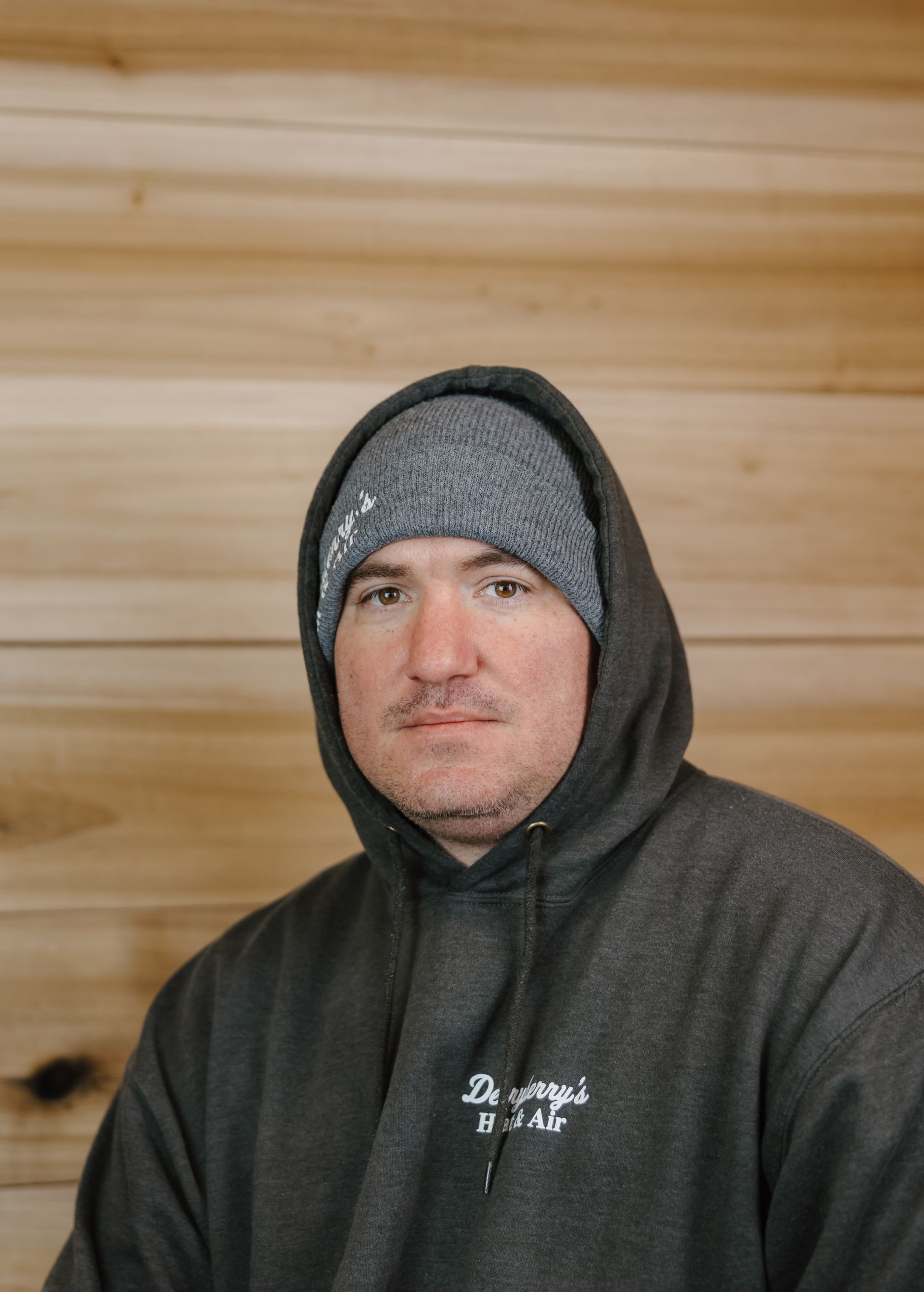 Man wearing a gray beanie and hooded sweatshirt against a wood-paneled wall, looking at the camera.