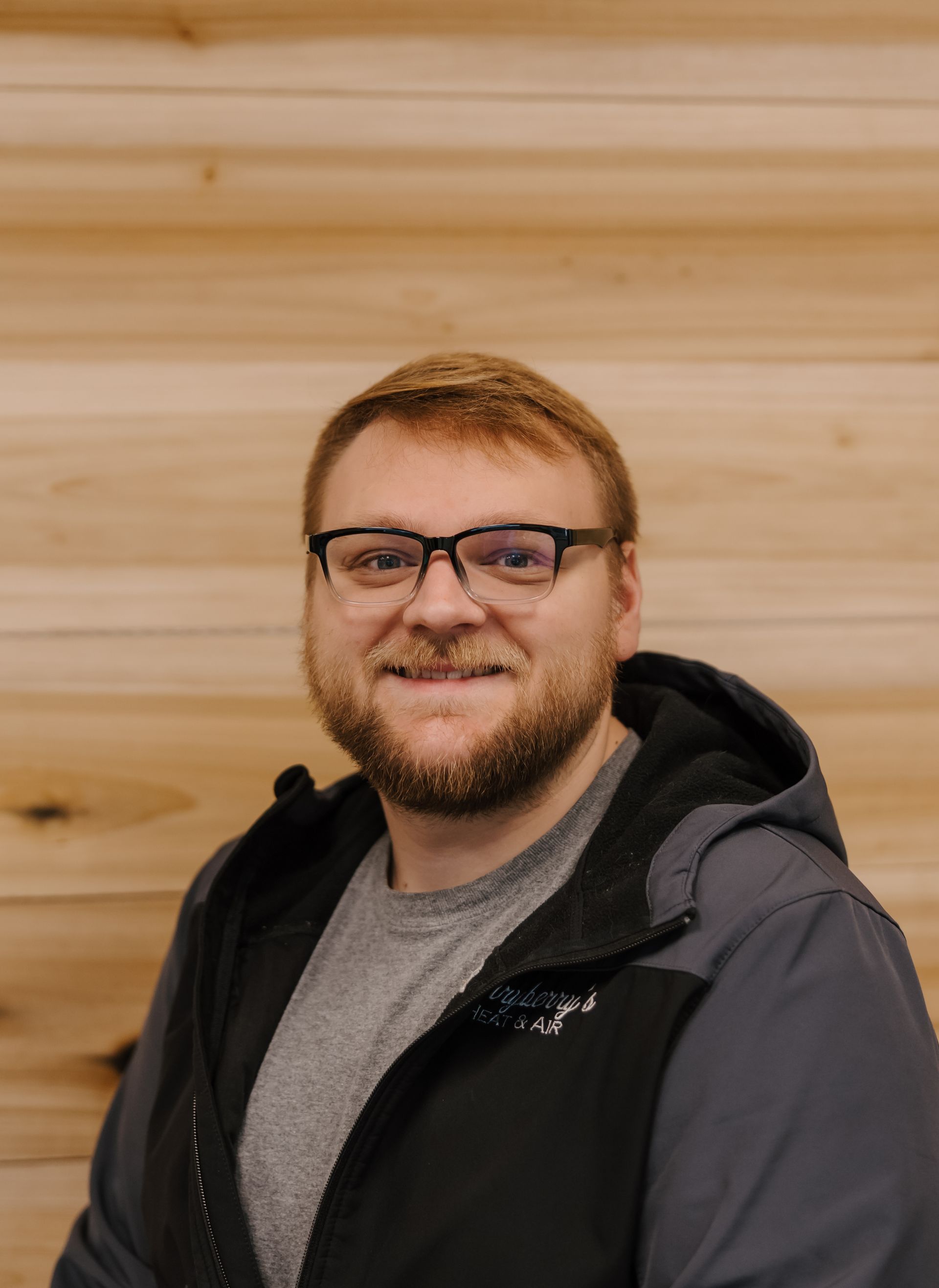 Man with glasses and beard, smiling. Wearing a gray shirt and black jacket. Standing in front of a wooden wall.