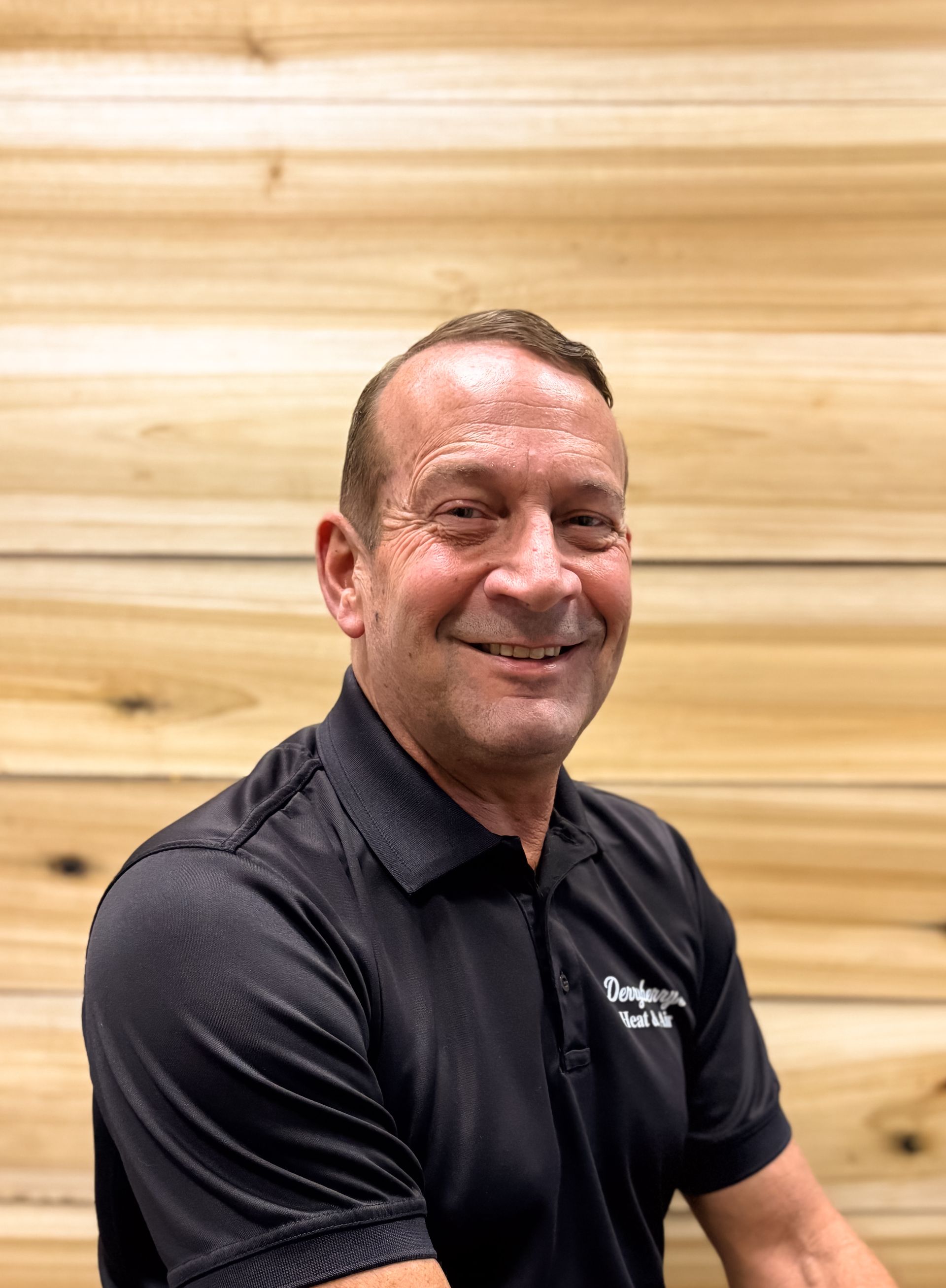 Man in black polo smiling in front of a wood-paneled wall.