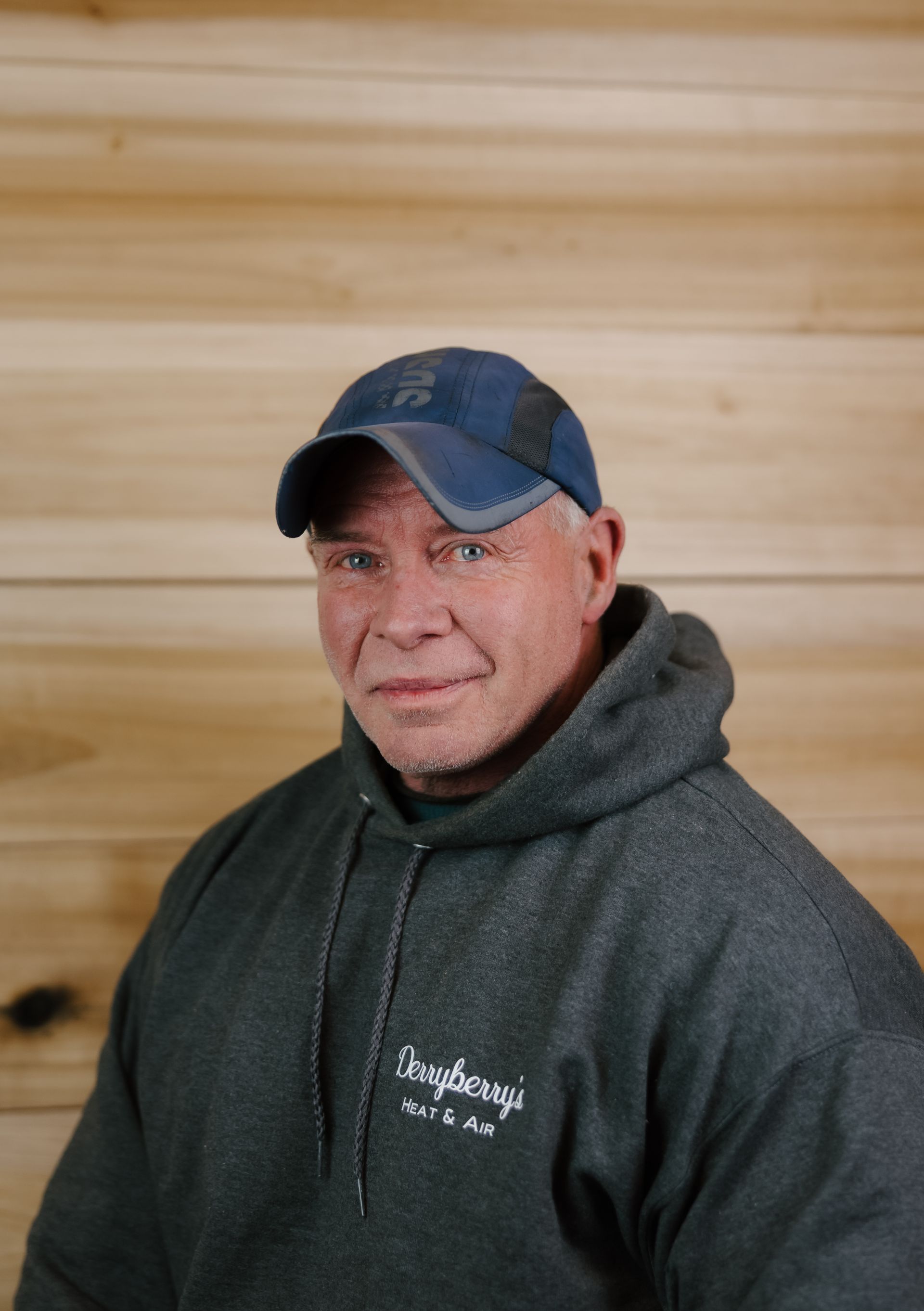 Man wearing a blue cap and gray hoodie, standing against a wooden wall.