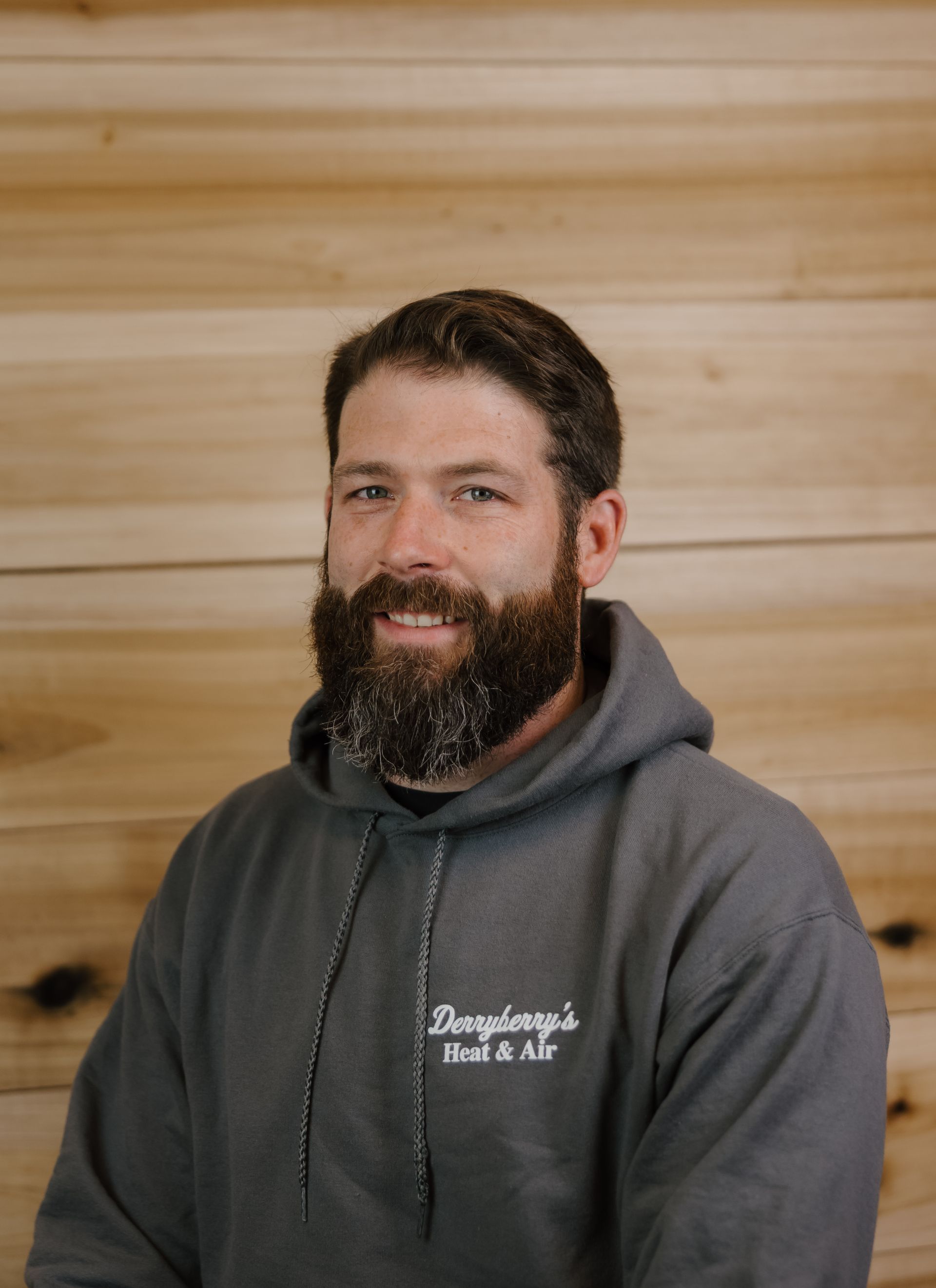 Man with dark hair and full beard wearing a gray hoodie in front of a wood-paneled wall.