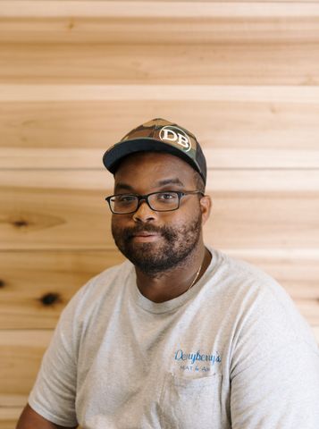 Man with a long beard and a hat wearing a gray hoodie in front of a wood panel background.
