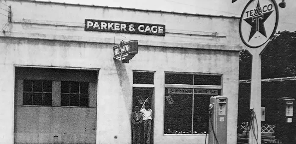 A black and white photo of a gas station named Parker & Cage, with a Texaco sign. Two people stand in the doorway.