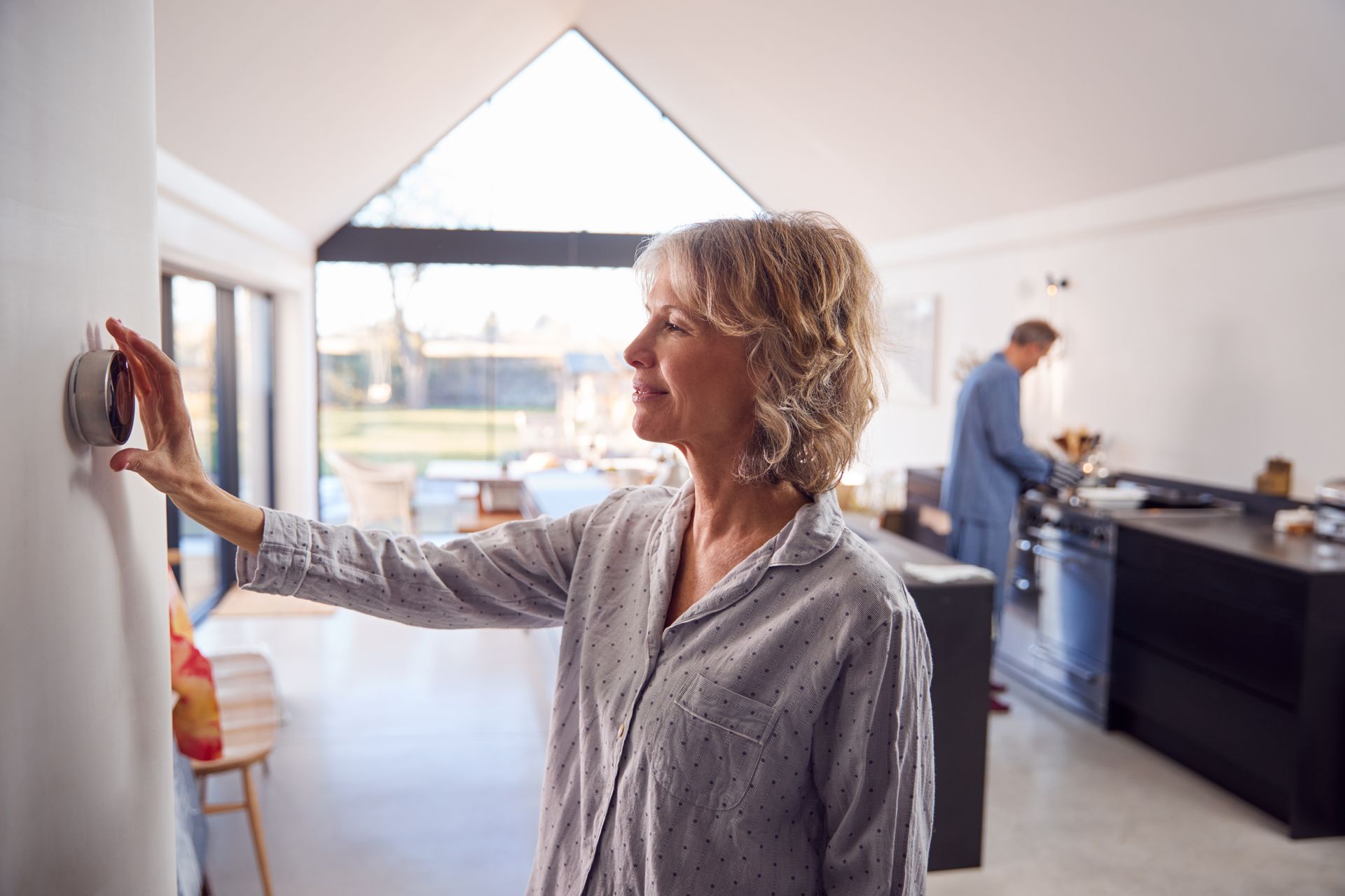 Woman adjusting a thermostat on a white wall in a modern kitchen. Man in background.