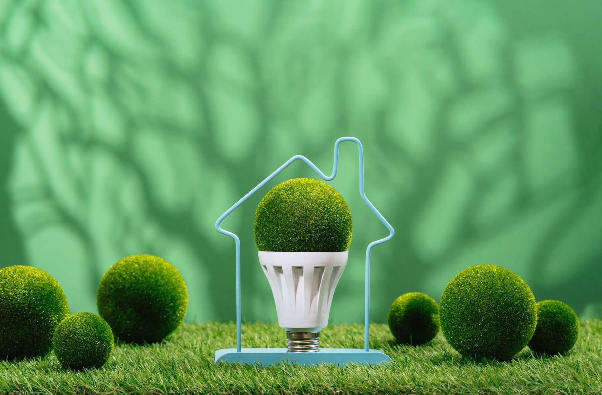 Green LED light bulb in a house frame, surrounded by green spheres on artificial grass, green background.