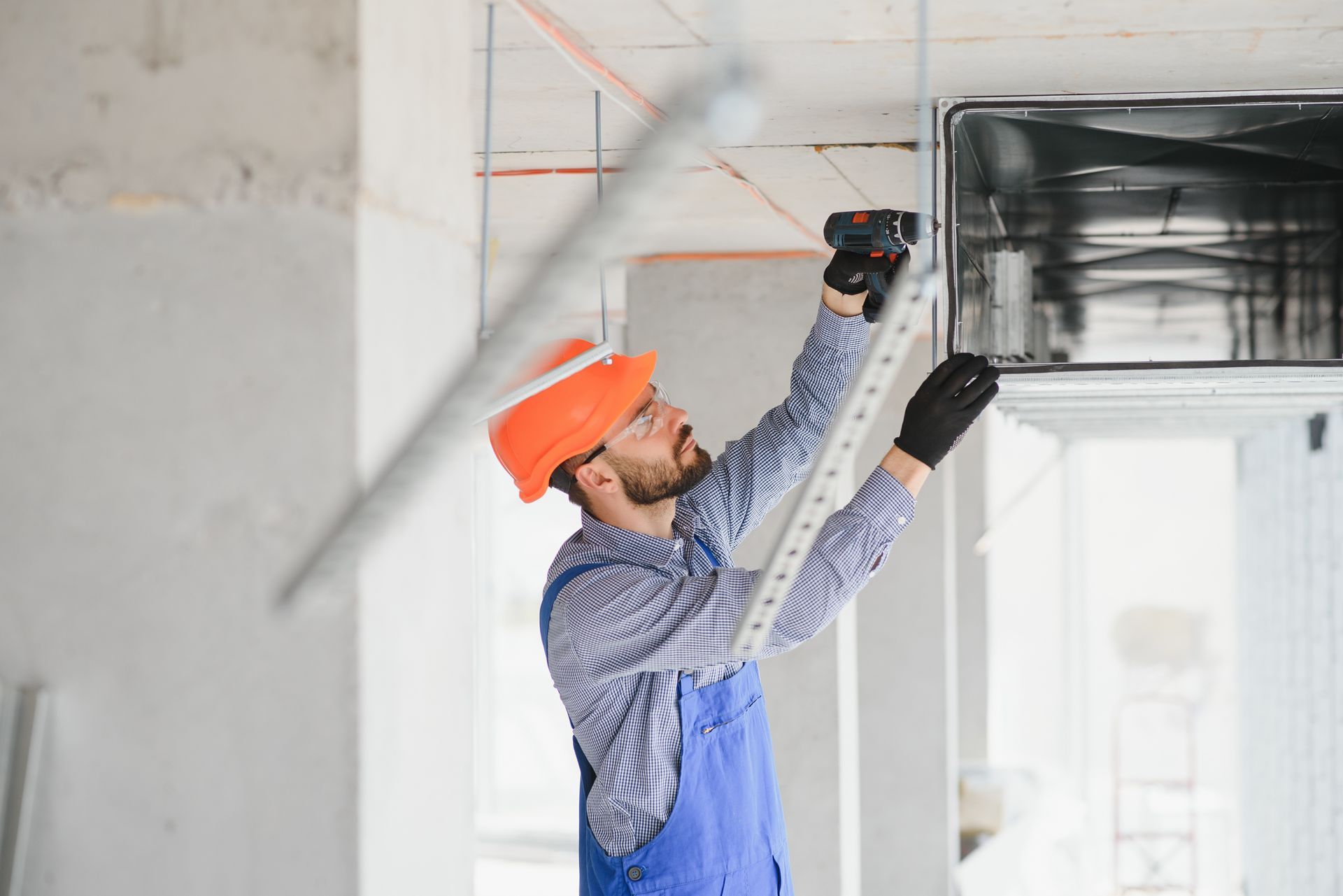 Construction worker in orange hard hat measures with a level, installing ventilation duct in a building.