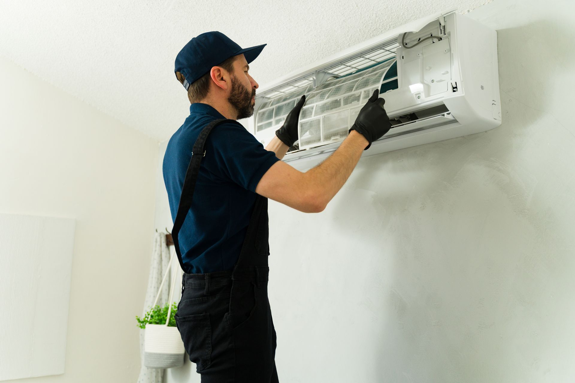 HVAC technician in black overalls and gloves cleaning an AC unit on a white wall.