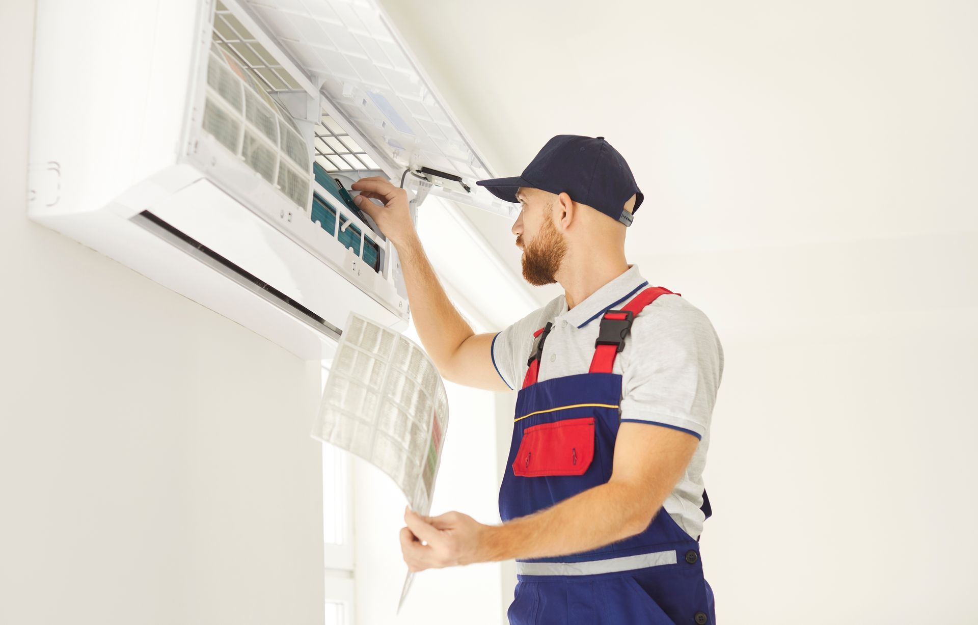 HVAC technician in overalls cleans an air conditioner unit, white walls.