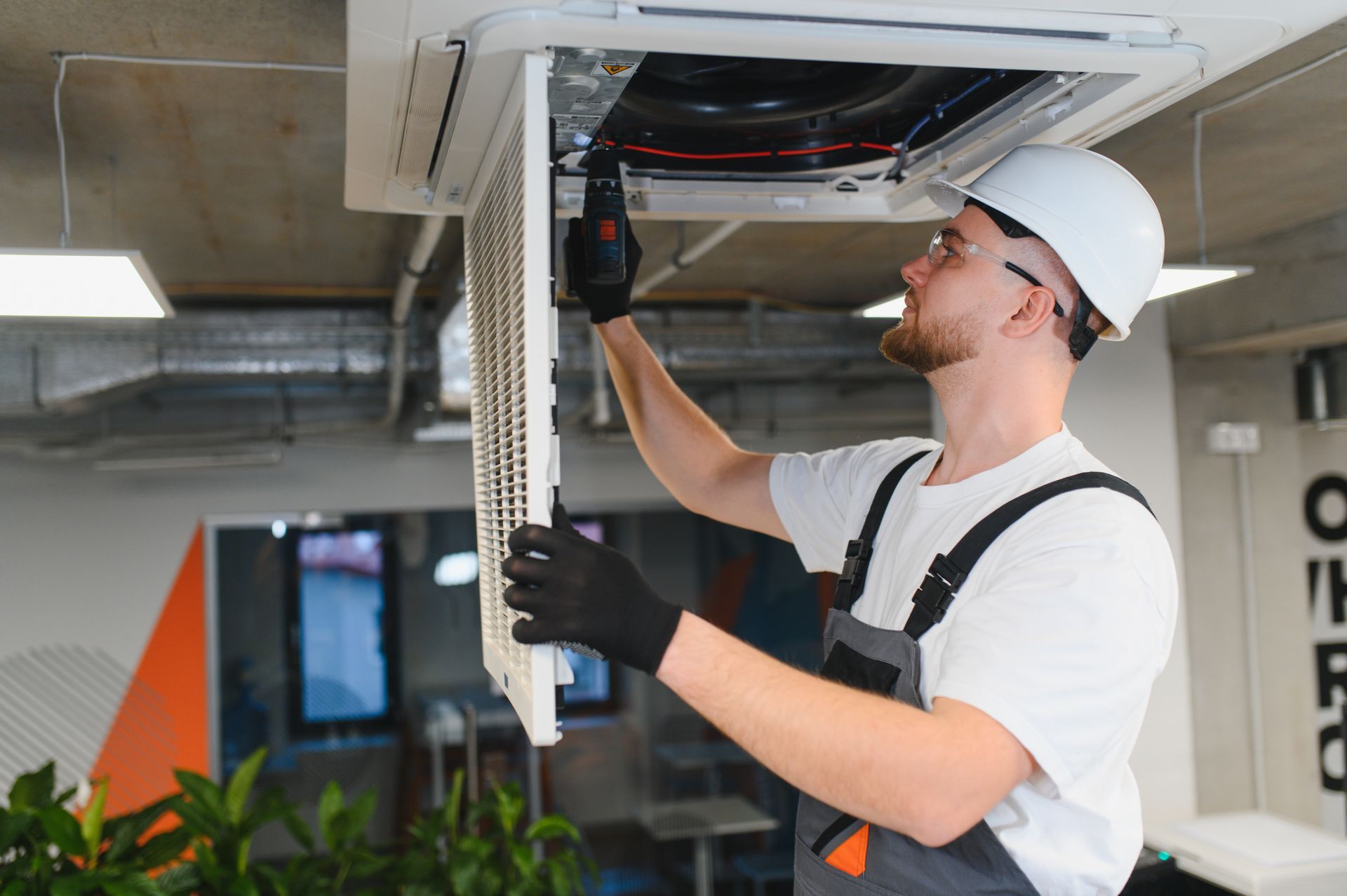 HVAC technician in white hardhat and gloves removes an air filter from a ceiling unit.