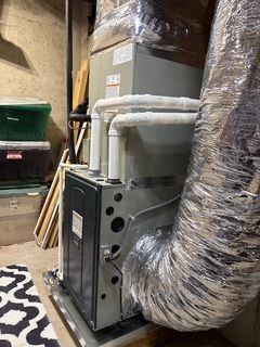 Furnace in a basement, with ductwork and nearby storage containers. Gray and silver colors dominate.