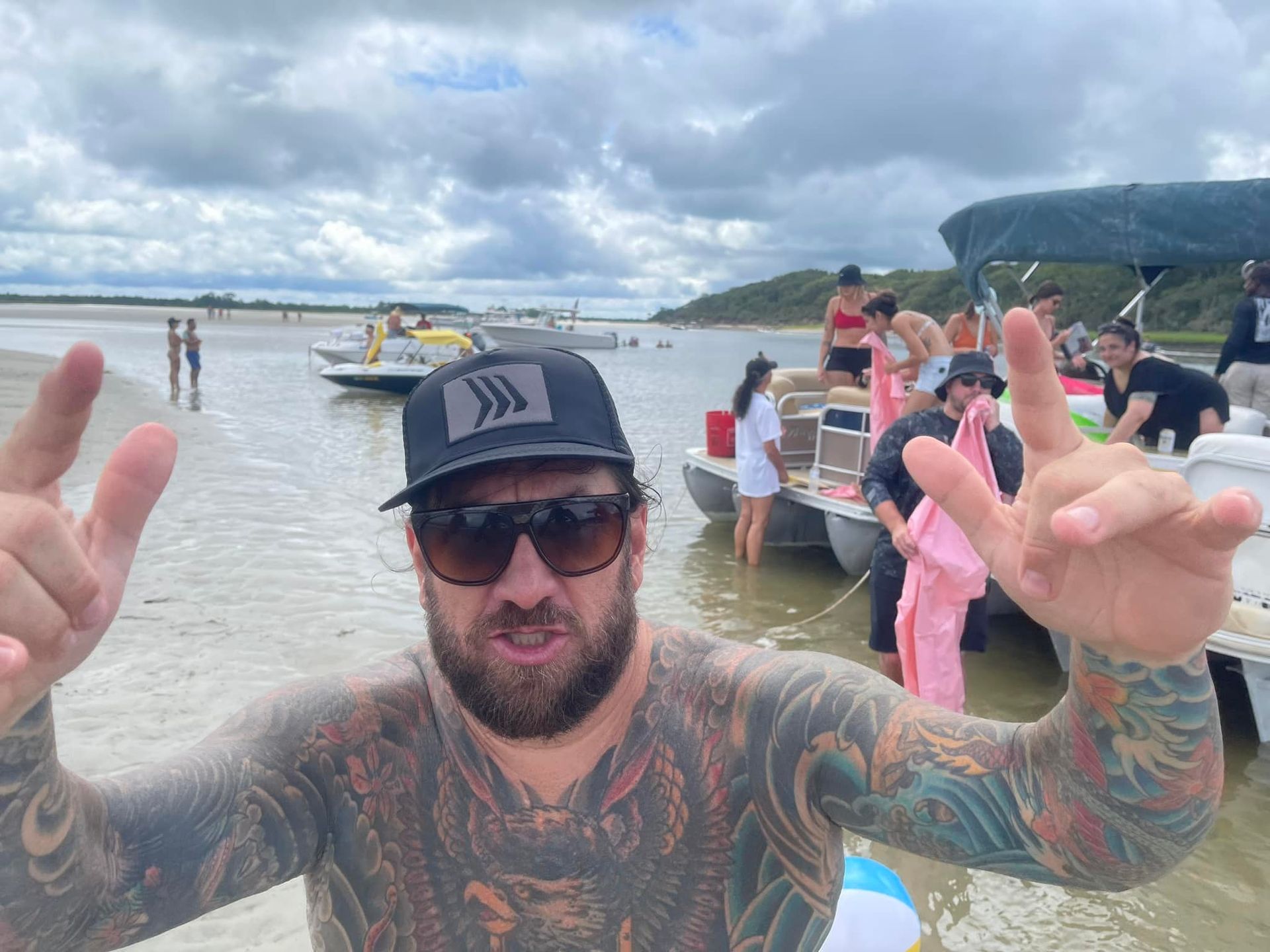 A man with a beard and tattoos is standing in the water with boats in the background.