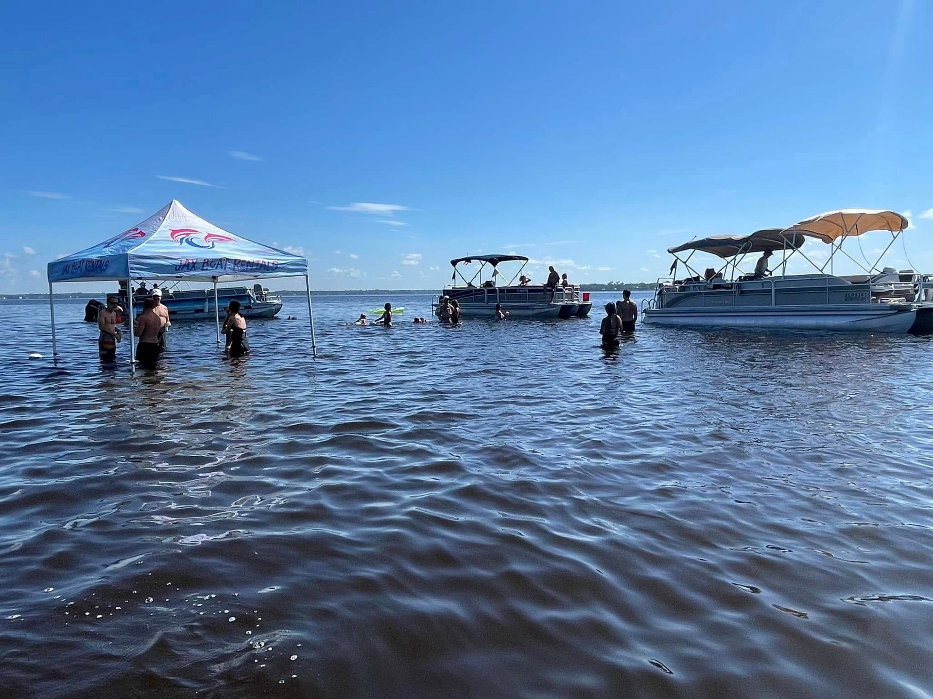 A group of people are standing in the water with boats in the background.