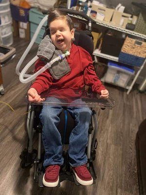 A young boy is sitting in a wheelchair with a ventilator attached to his neck.
