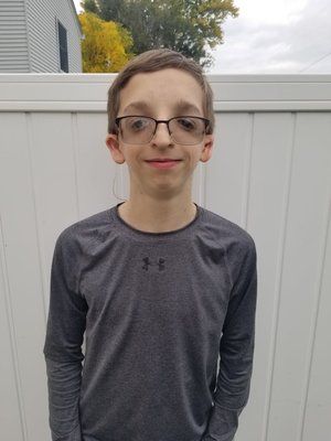 A young boy wearing glasses and a gray shirt is standing in front of a white fence.