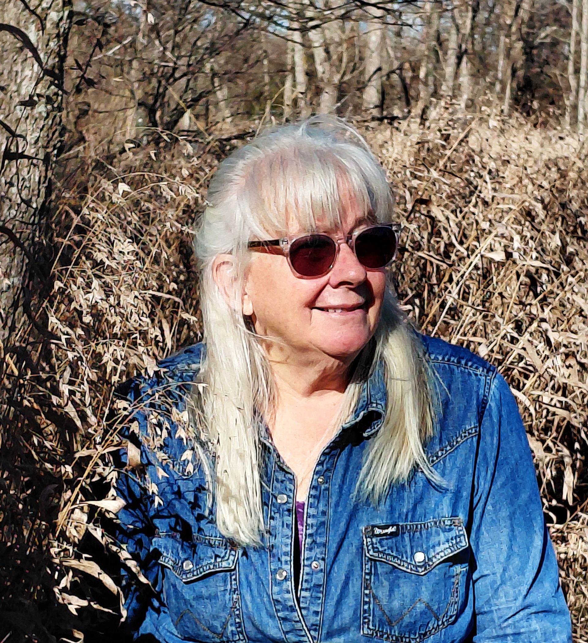 Woman with white hair and sunglasses smiles, wearing a denim shirt, outdoors, with a backdrop of dried foliage.
