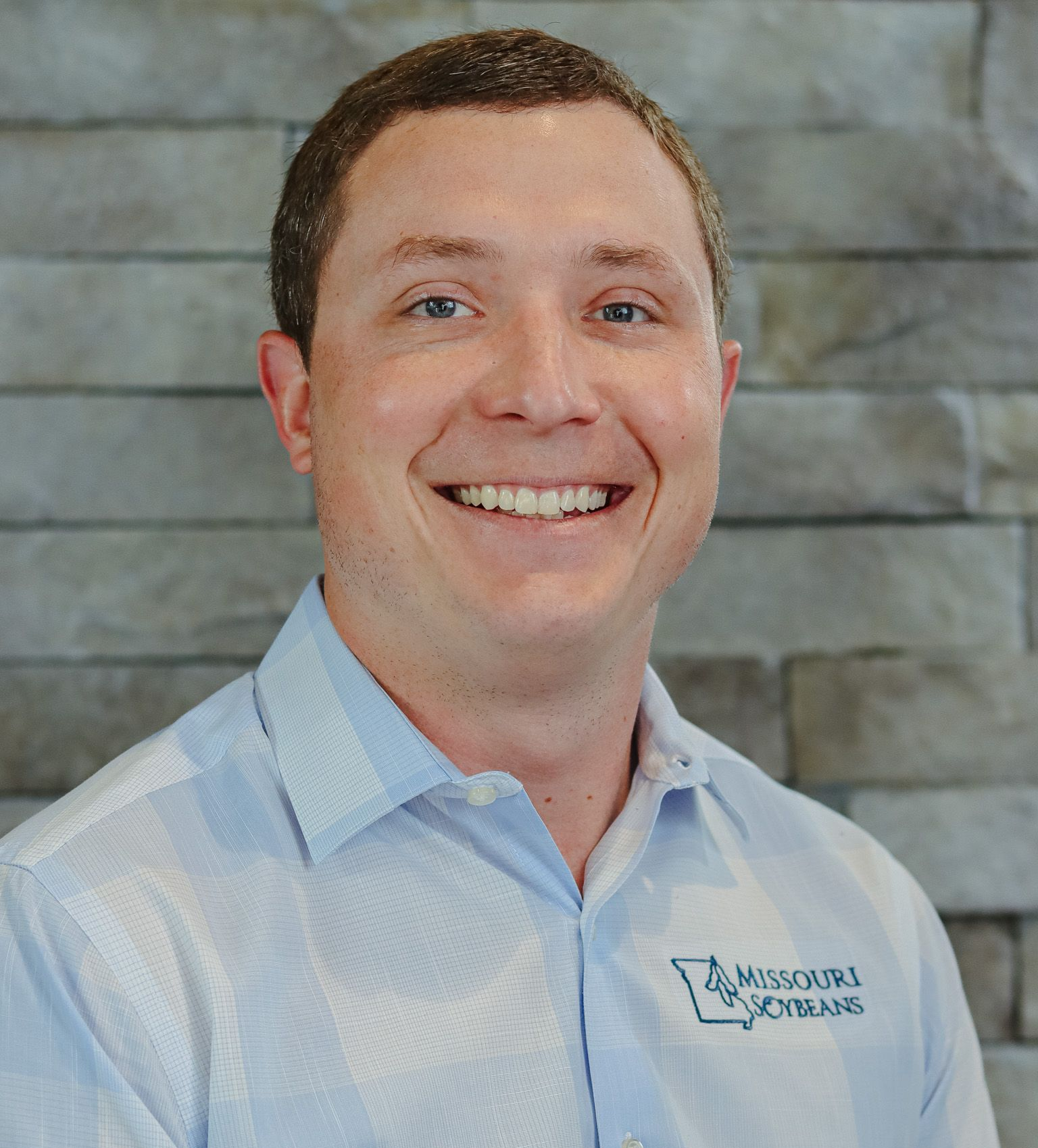 Man in light blue shirt smiling, Missouri Soybean logo on shirt, stone wall background.