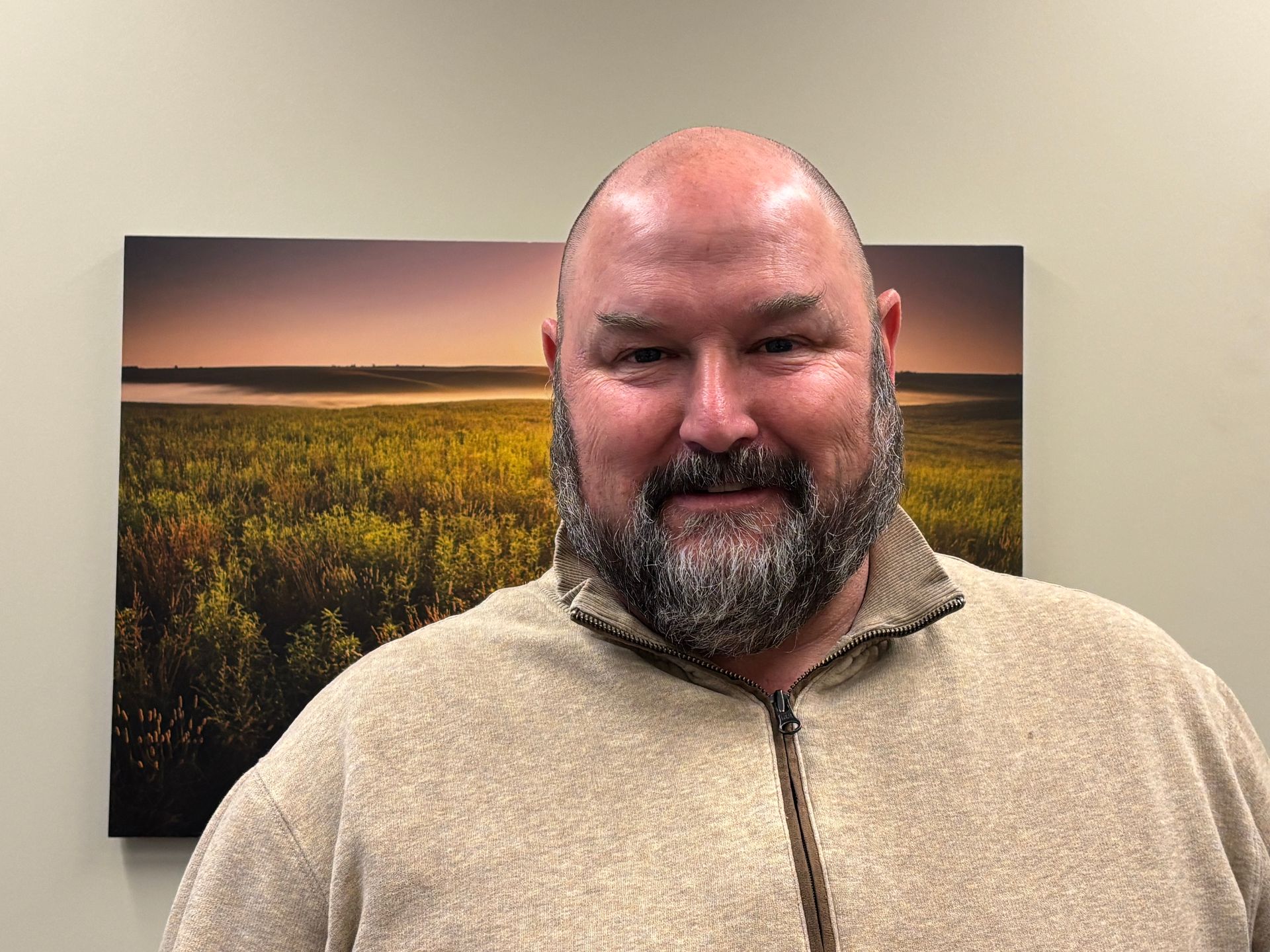 Man with beard and bald head wearing a tan zip-up sweater smiles in front of a landscape photo.