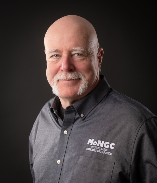 Bald man with a white mustache wearing a grey shirt with 